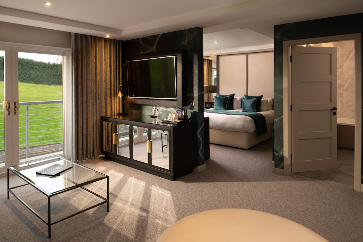Une chambre d’hôtel moderne avec une table basse en verre, un lit agrémenté de coussins turquoise, un téléviseur fixé au-dessus d’une console sombre, et une porte donnant sur un balcon avec vue sur un paysage verdoyant.