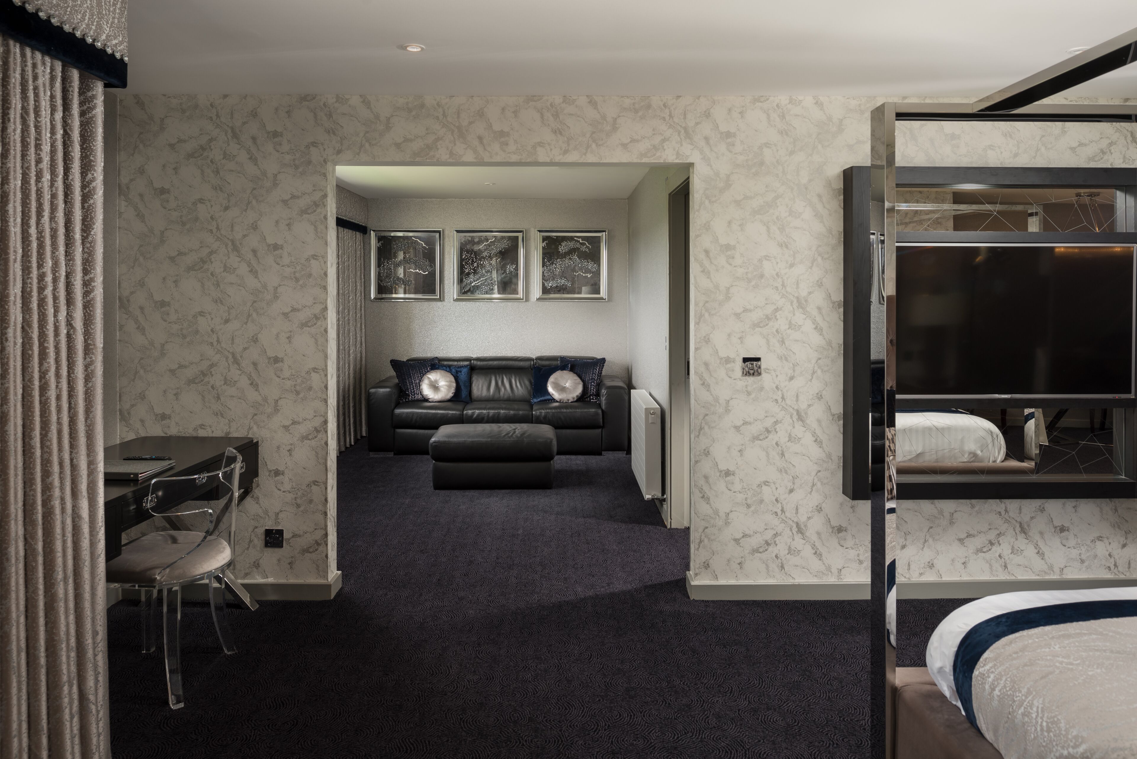 Habitación de hotel con alfombra oscura, un escritorio con silla, sofá con cojines, cama con cabecera azul y televisor montado en la pared.