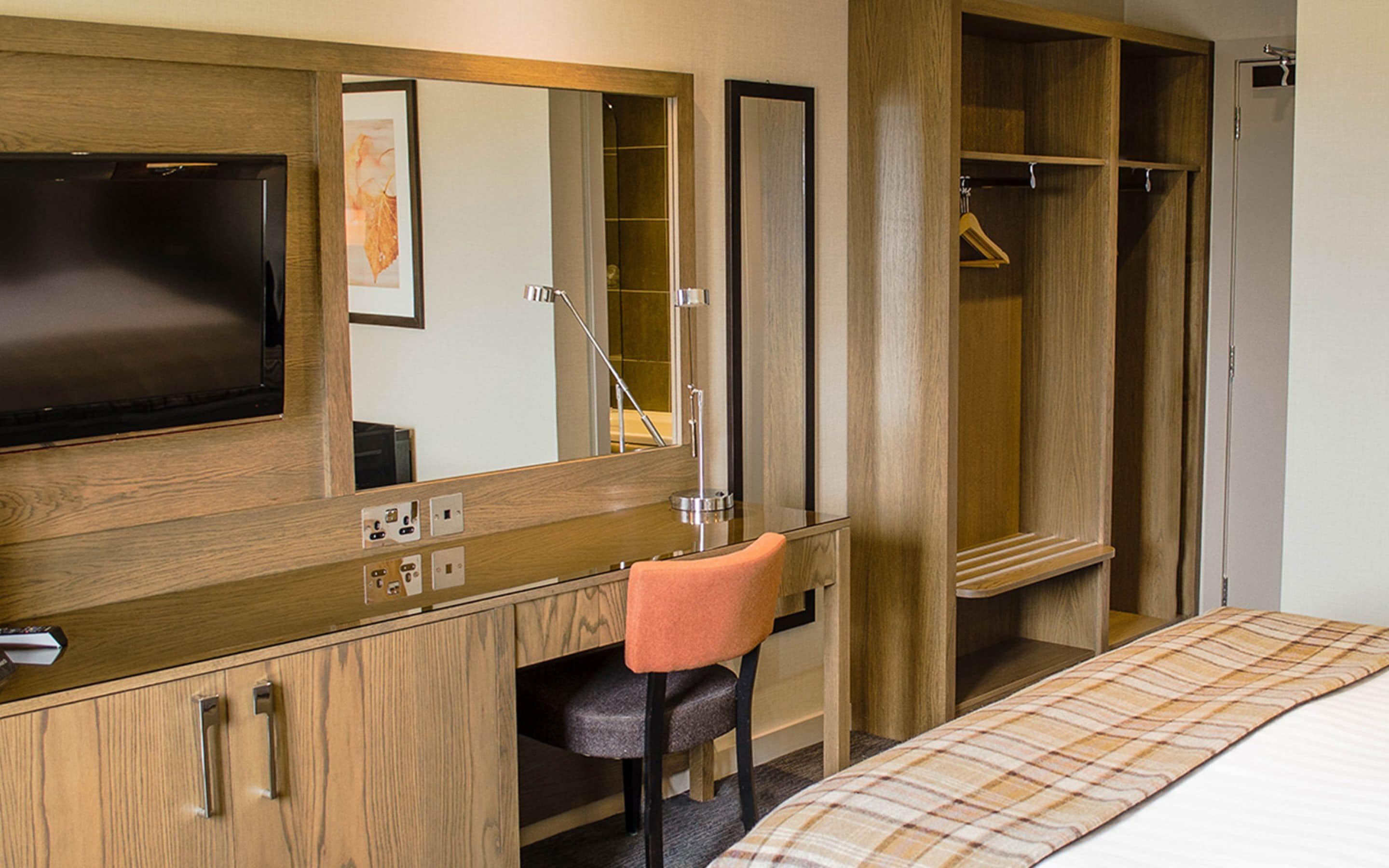 Une chambre d’hôtel avec un bureau en bois, une chaise, un lit recouvert d’une couverture à carreaux, un grand miroir, une télévision et un dressing.
