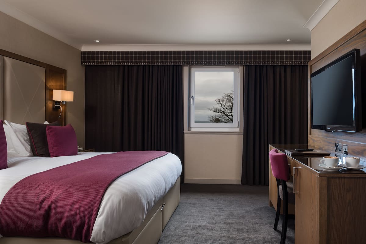 Une chambre d’hôtel comprenant un lit, un bureau avec une chaise, une télévision et une fenêtre avec des rideaux.