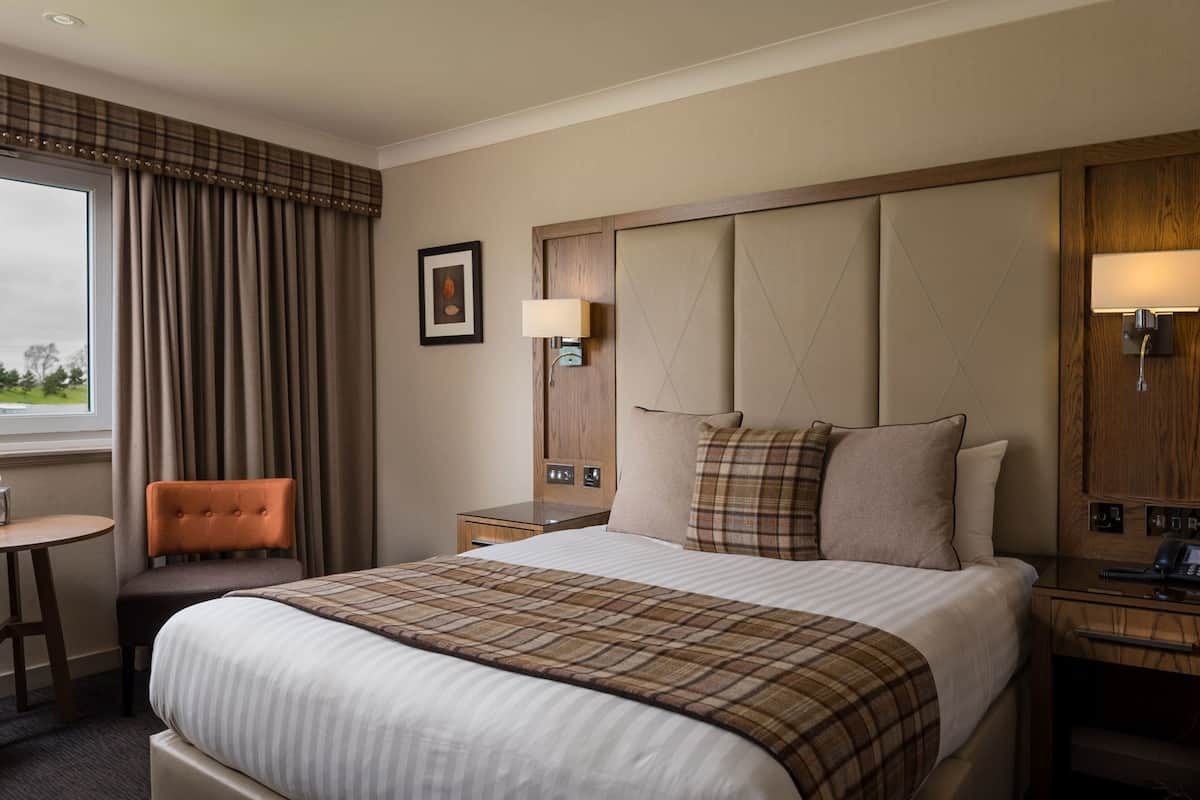 Une chambre d’hôtel avec un lit, une chaise, une table de chevet, une lampe et une fenêtre avec des rideaux.