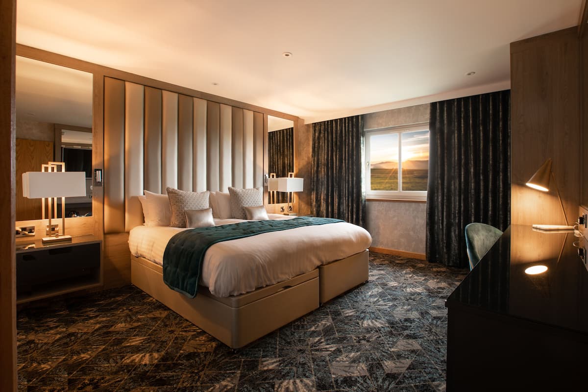 Une chambre d’hôtel moderne avec un grand lit, deux tables de chevet avec des lampes, un bureau et une vue sur le coucher de soleil par la fenêtre.