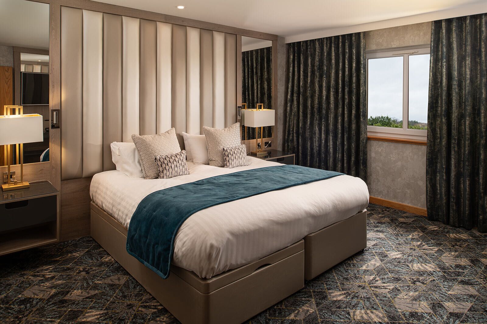 Une chambre d’hôtel moderne avec un grand lit, une table de chevet avec une lampe, une commode et une grande fenêtre avec des rideaux.
