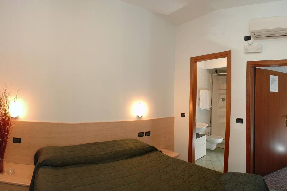 A hotel room with a bed, a wooden wardrobe, a bedside table with a lamp, and a bathroom visible through an open door.
