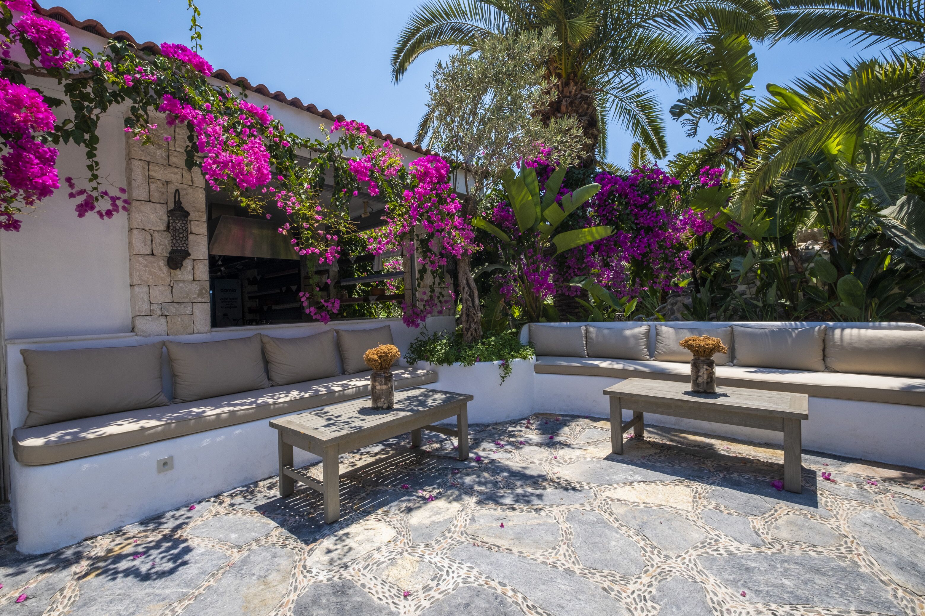 An outdoor seating area with stone flooring, cushioned benches, and a pergola covered with flowering vines.