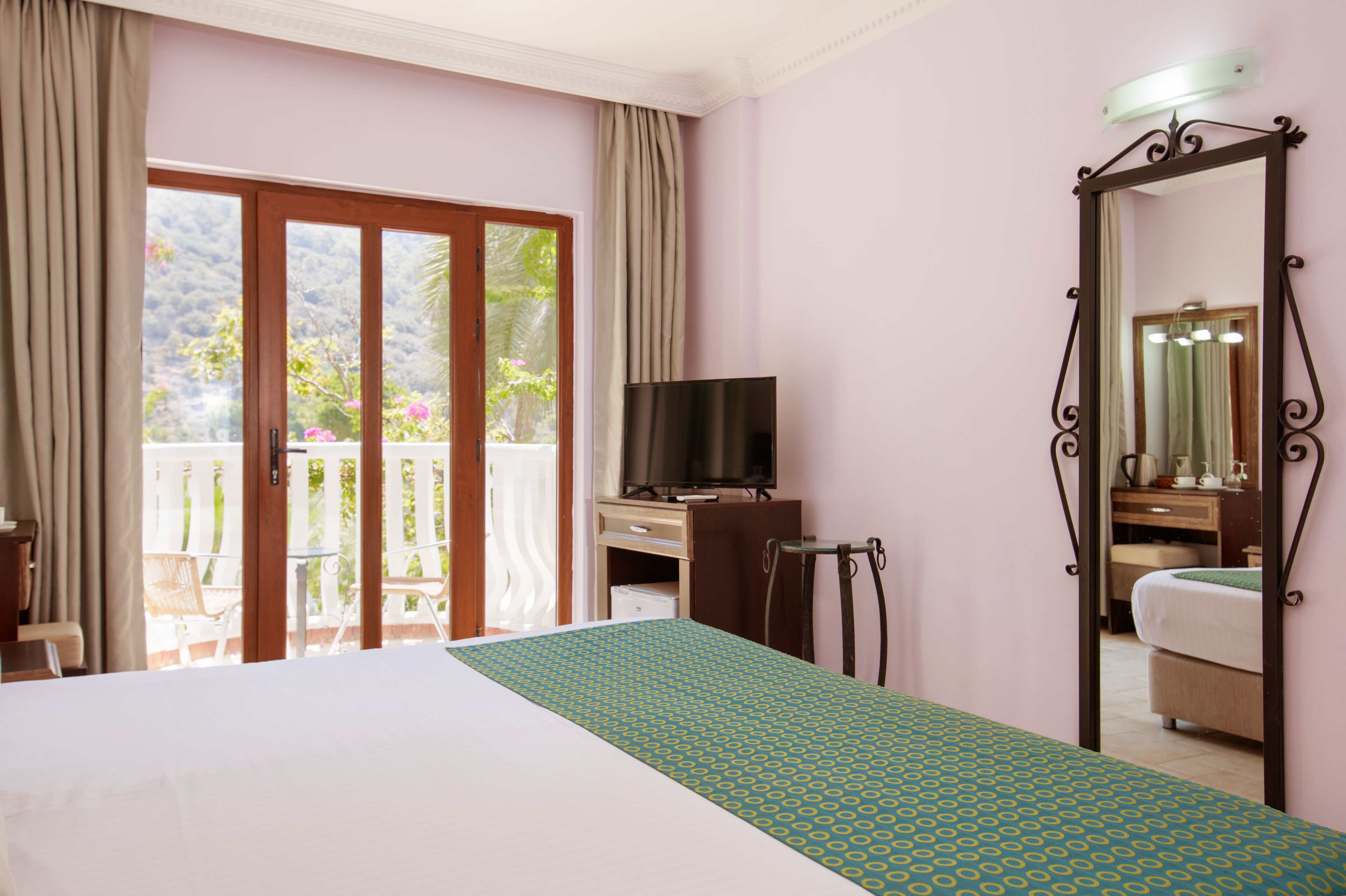 Une chambre d’hôtel avec un lit, un bureau avec une télévision, un miroir et un balcon donnant sur un paysage verdoyant.