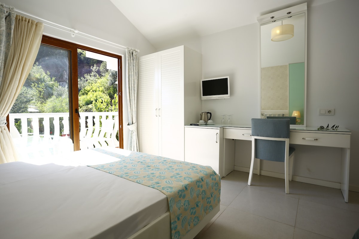 A bedroom with a bed, a desk, a chair, a TV, and a balcony with curtains.