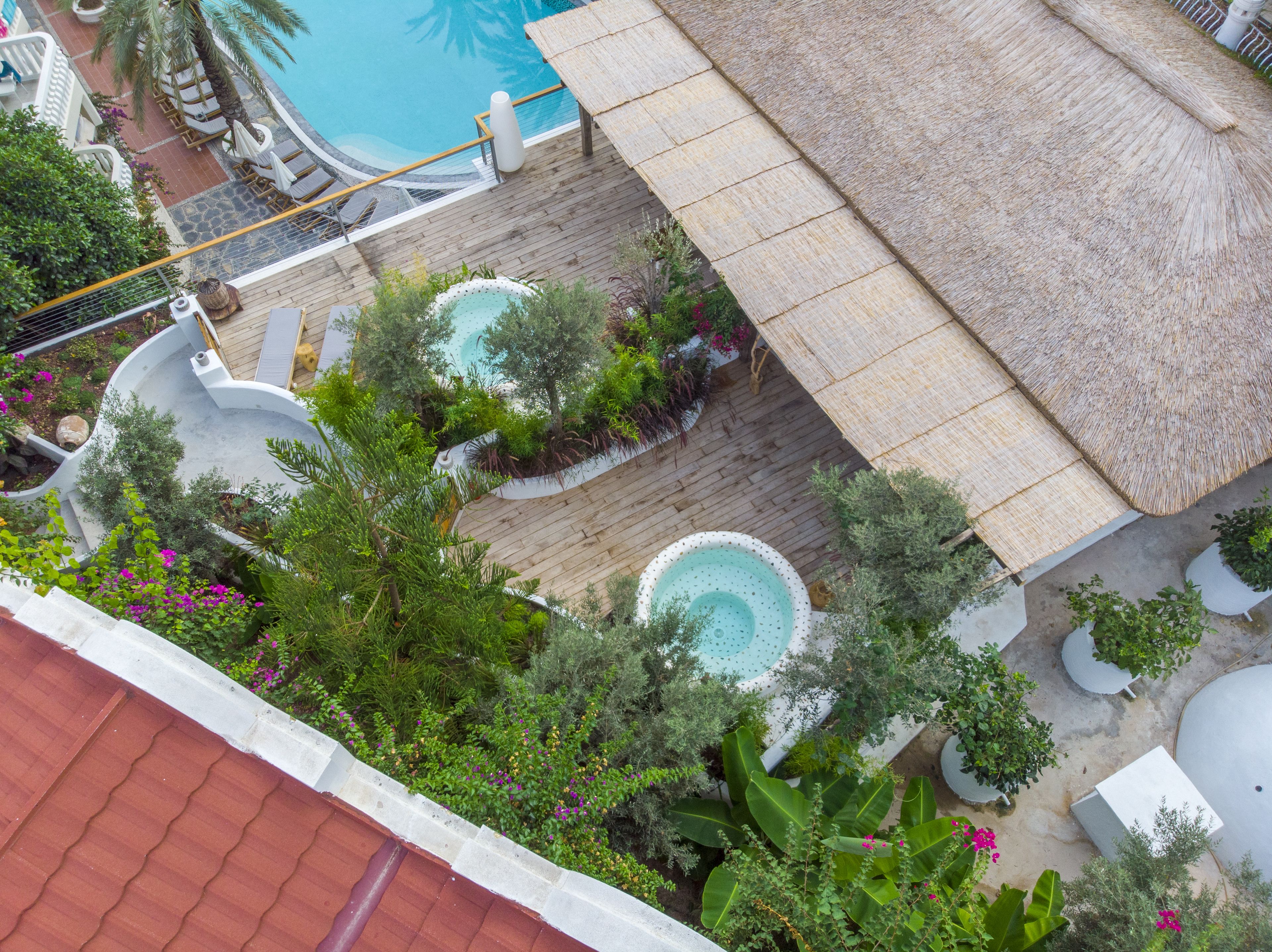 A rooftop pool area with multiple water features, including a central pool and two smaller hot tubs, surrounded by lush greenery and a thatched roof structure.