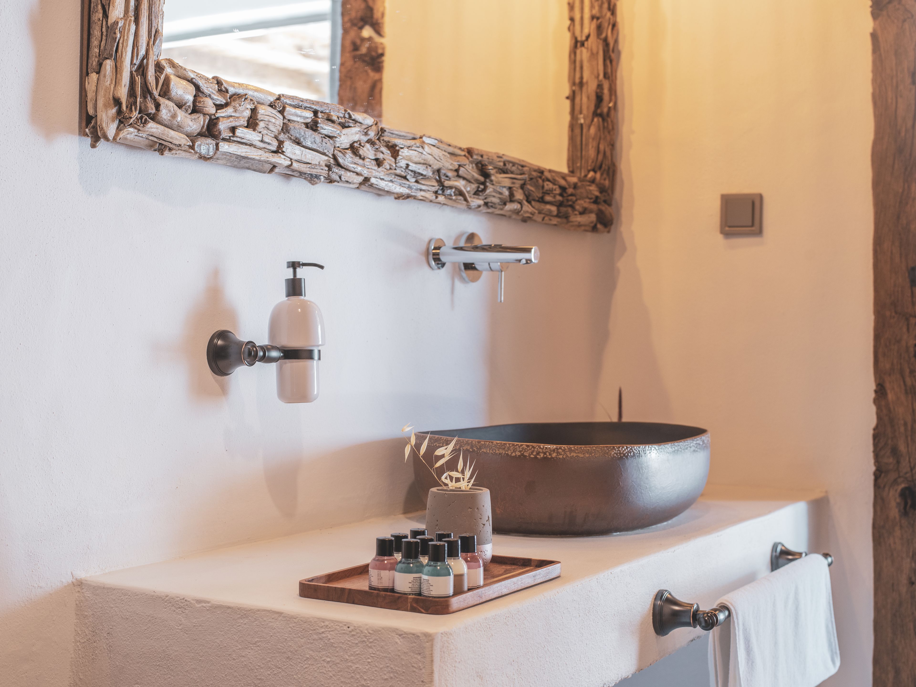 A bathroom with a large basin, a wall-mounted soap dispenser, a towel rack, and a mirror with a rustic frame.