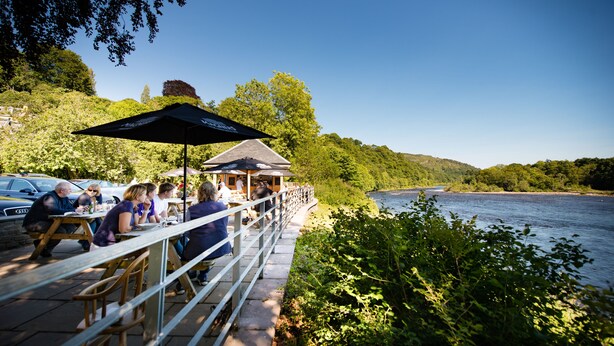 A riverside dining area with outdoor seating and umbrellas, overlooking a river. - Atholl Arms Hotel (Dunkeld)