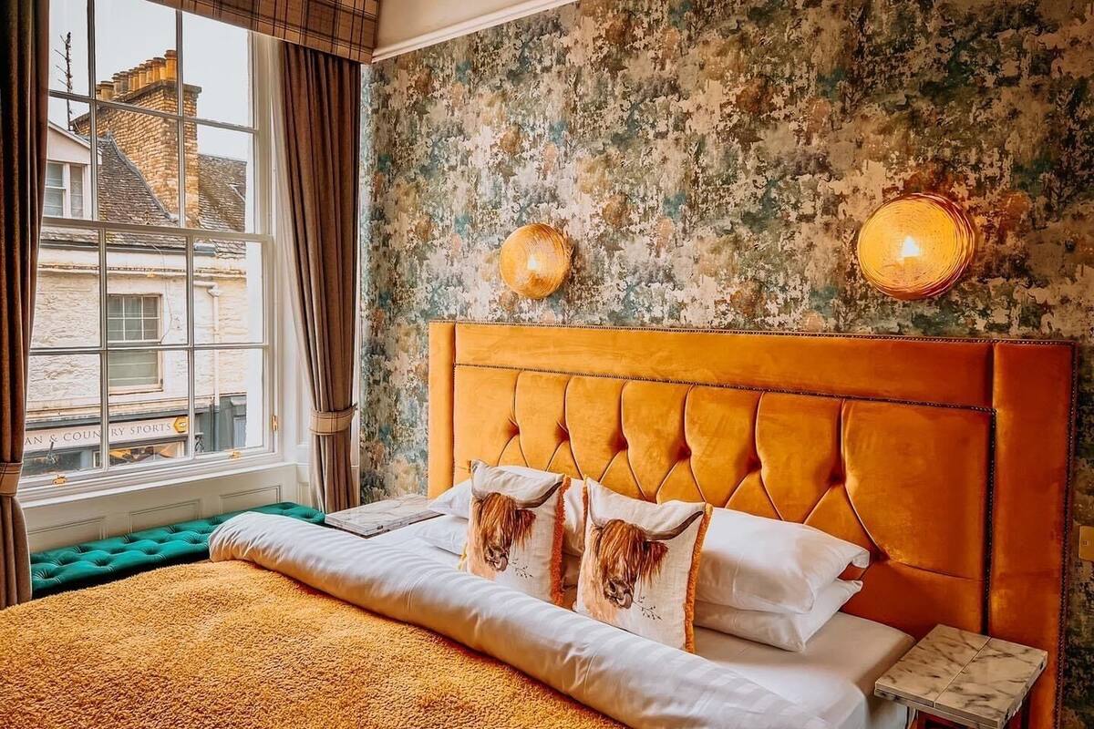 A bedroom with a large bed, a green upholstered bench, a rustic wall, and a window with curtains.