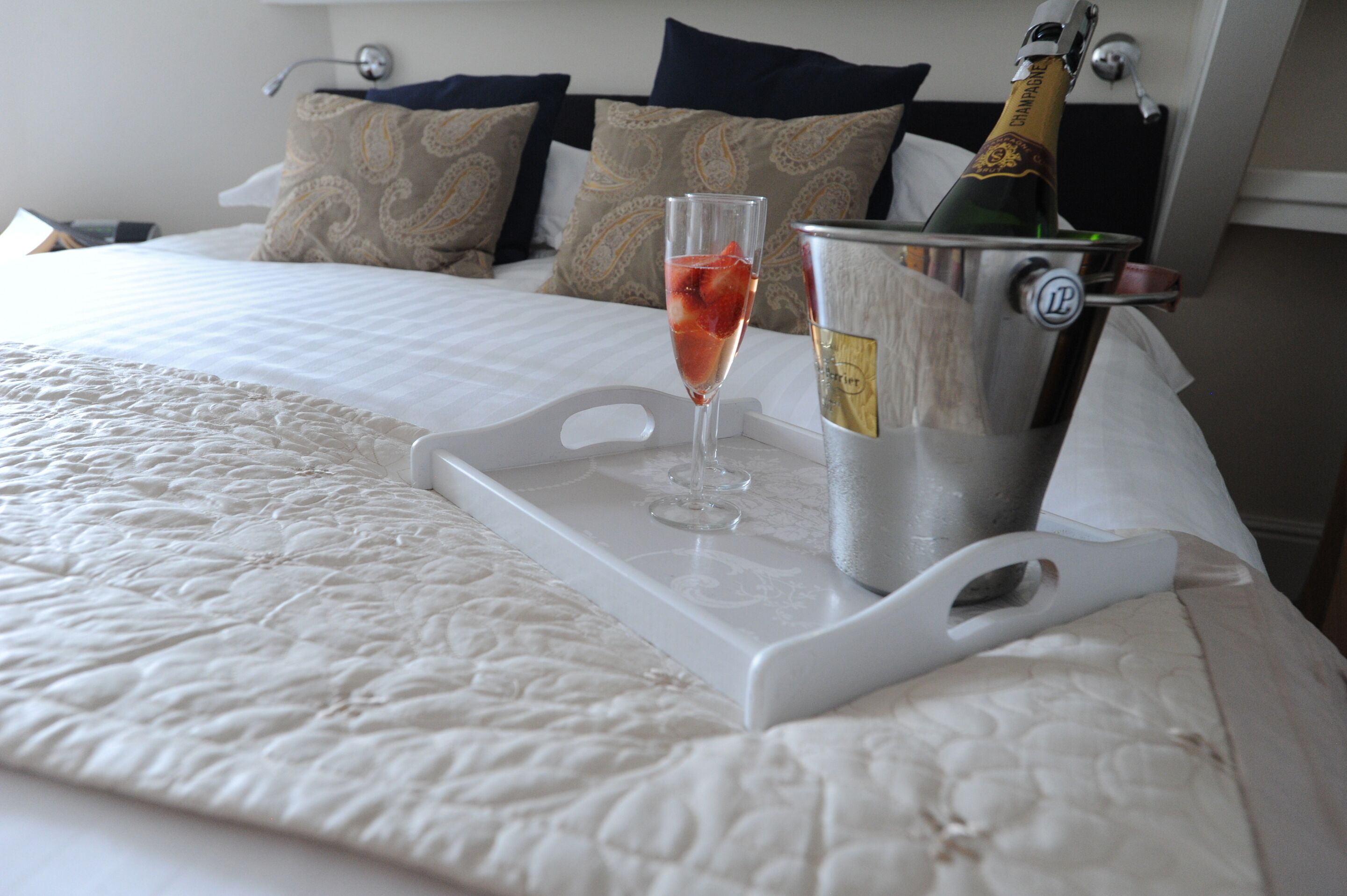 A bed with a white quilt, a champagne bottle in a metal bucket, and a glass of champagne on a tray.