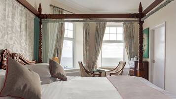 A four-poster bed with a view of the sea, a small table, and a chair in a room with a wooden ceiling and curtains.