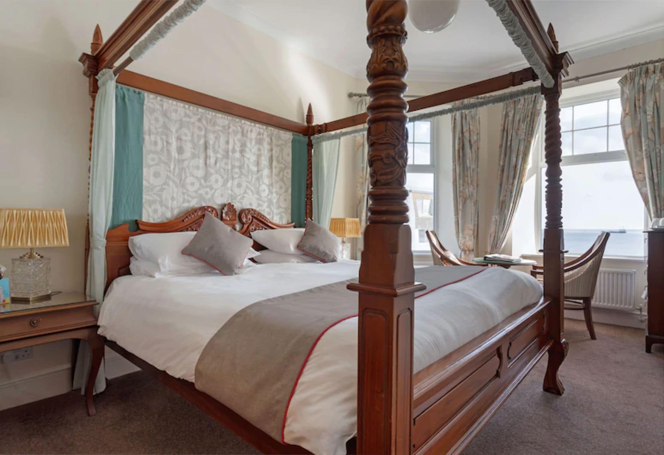 A four-poster bed with a wooden frame, a bedside table with a lamp, and a view of the sea through a window.