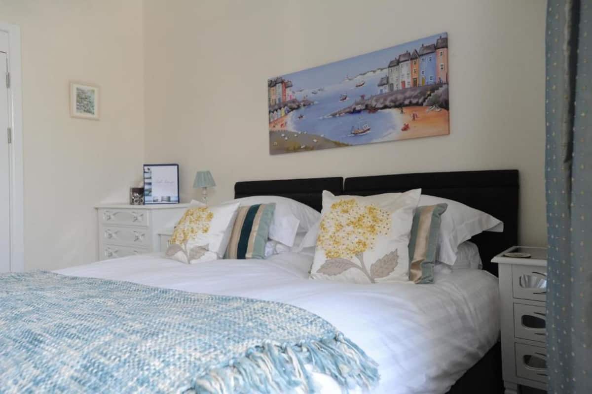 A neatly made bed with a blue and white blanket, a painting of a coastal scene, and a small bedside table with a lamp.