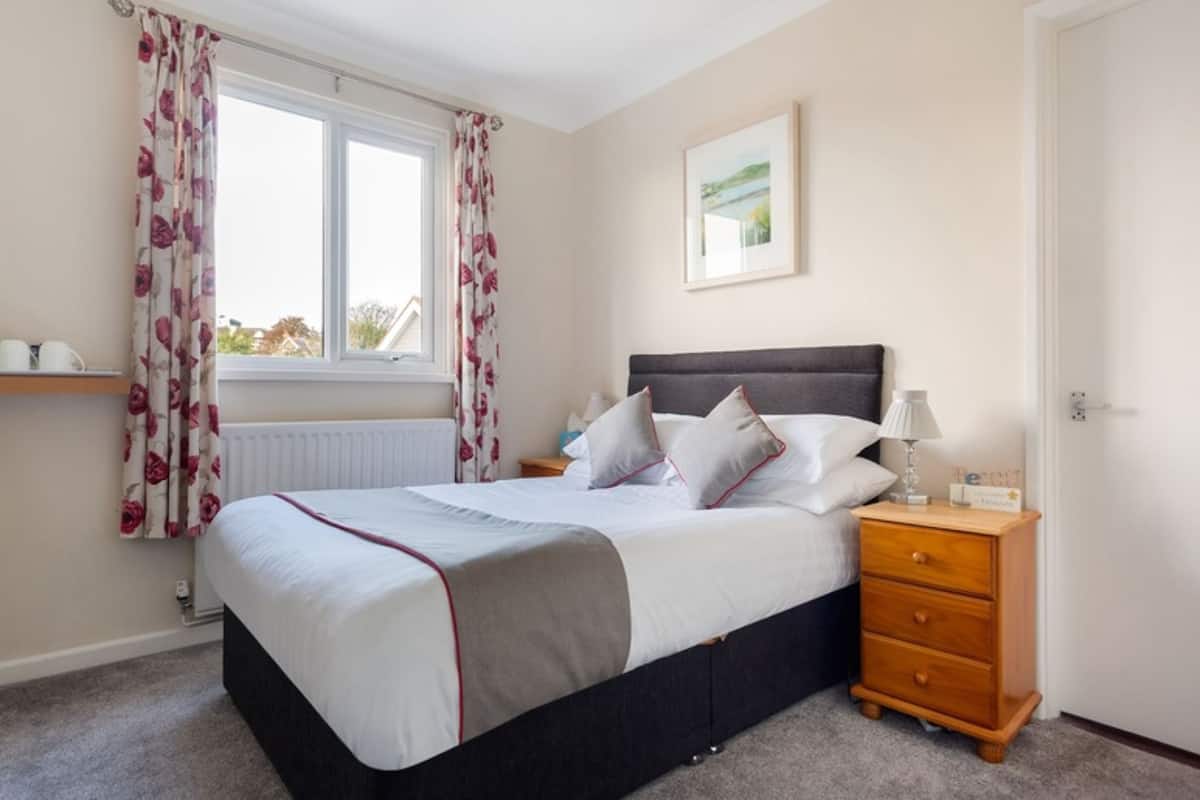 A bedroom with a bed, a nightstand, a window with curtains, a radiator, and a framed picture on the wall.