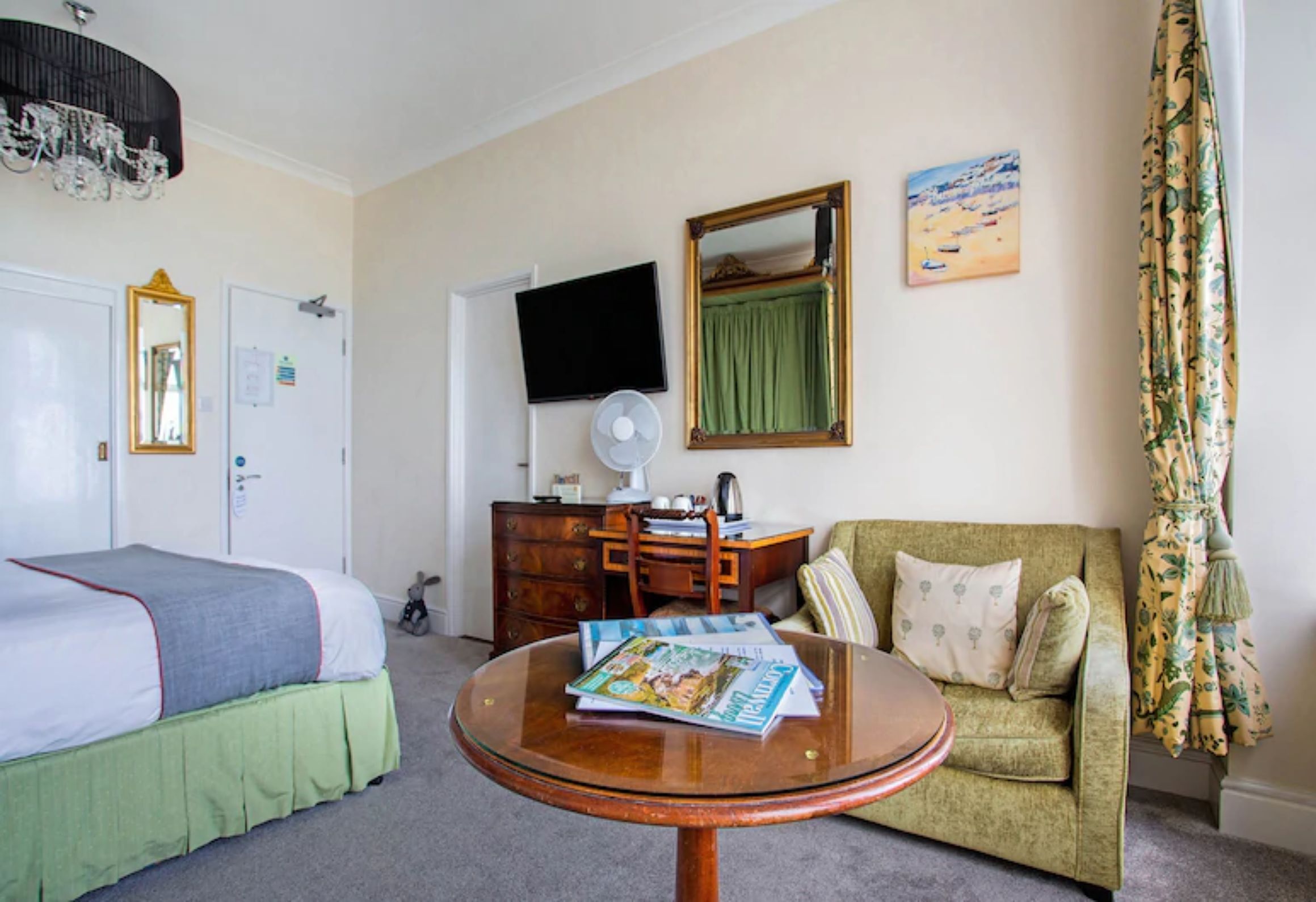 Une chambre d’hôtel avec un lit, une table ronde avec des magazines, un canapé vert, une commode, un téléviseur et un miroir.