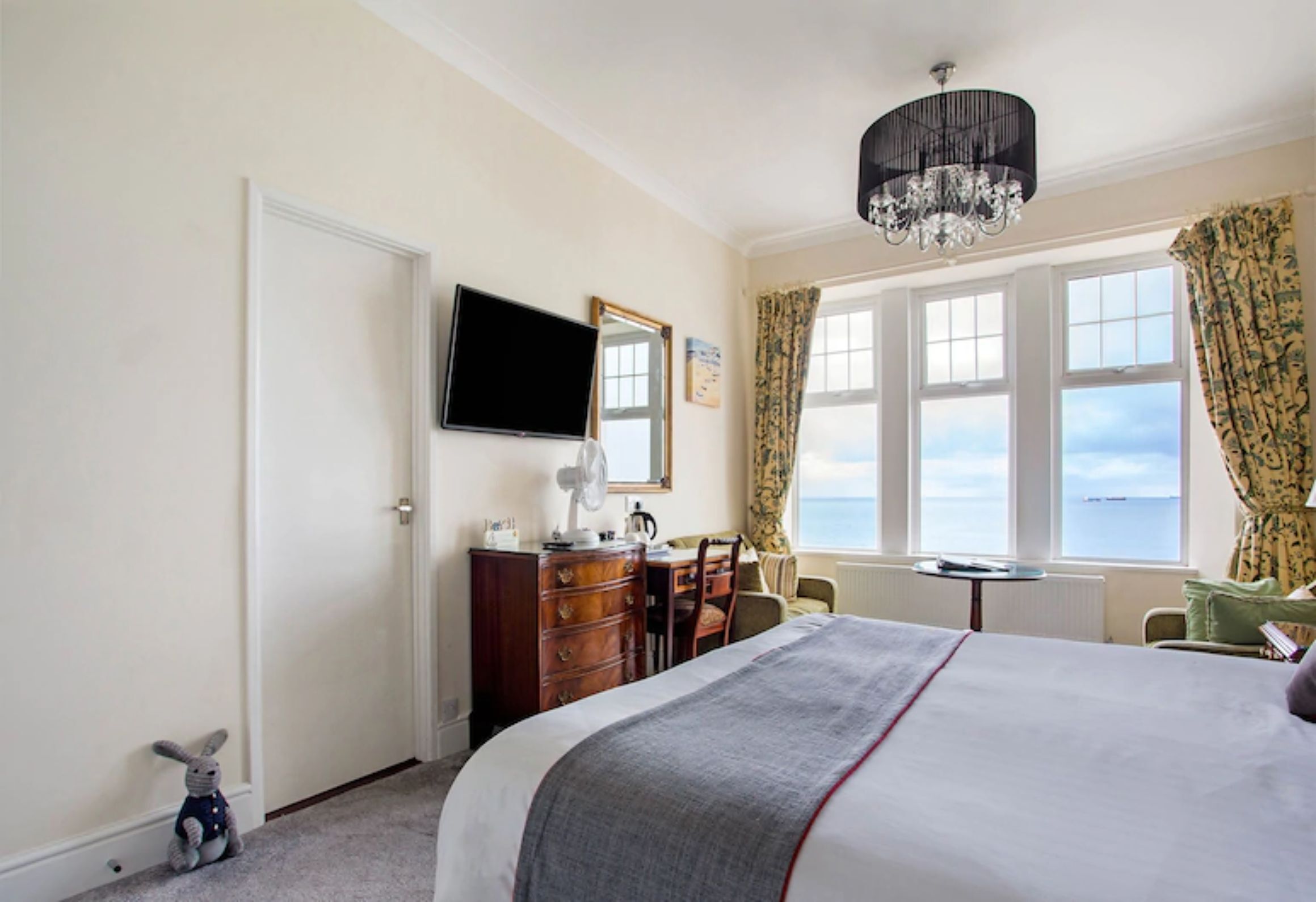 A hotel room with a bed, a dresser, a television, a desk, a chair, a small table, a window with curtains, and a view of the sea.