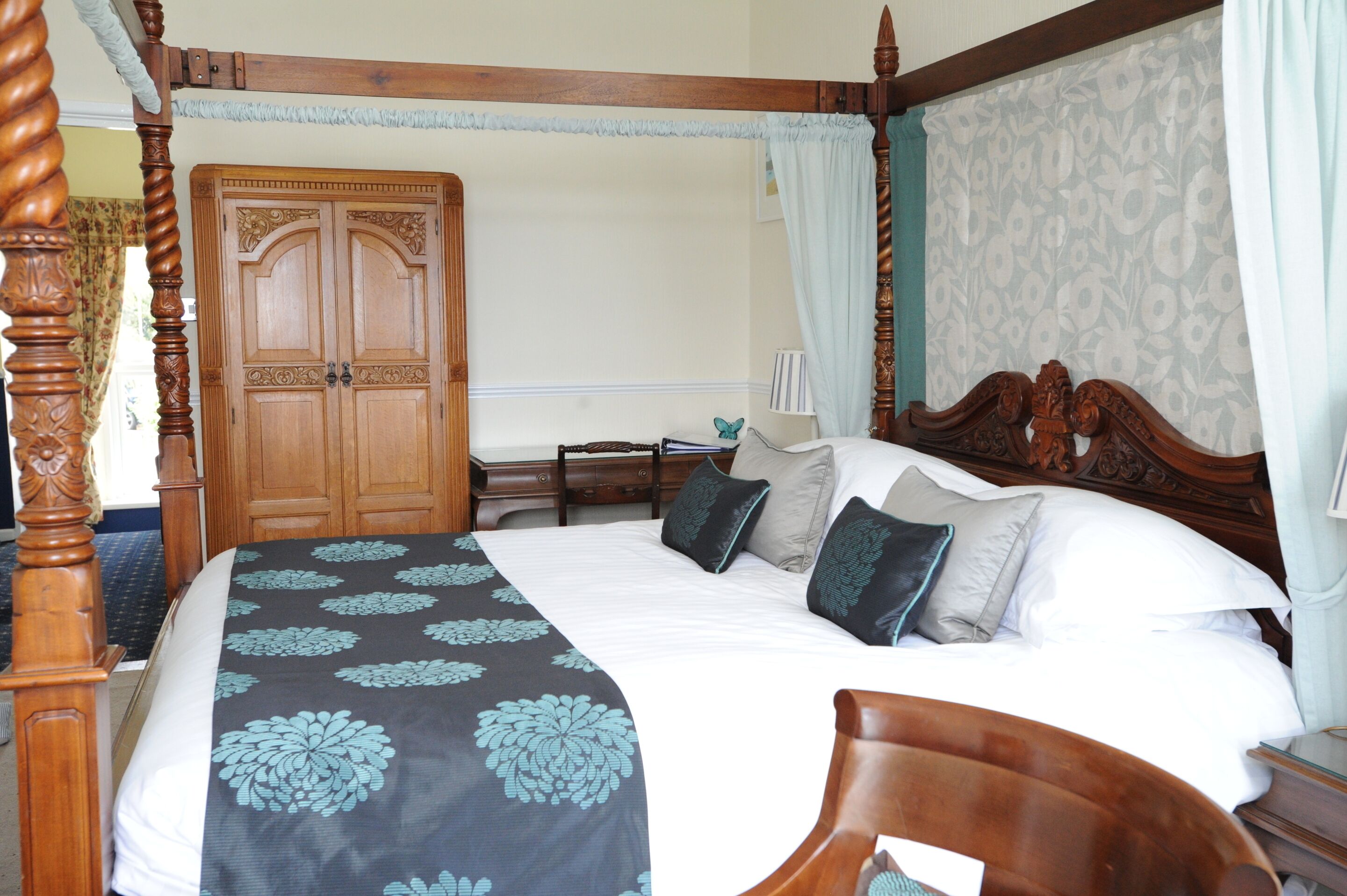 A four-poster bed with a floral bedspread, a wooden wardrobe, and a nightstand with a lamp.