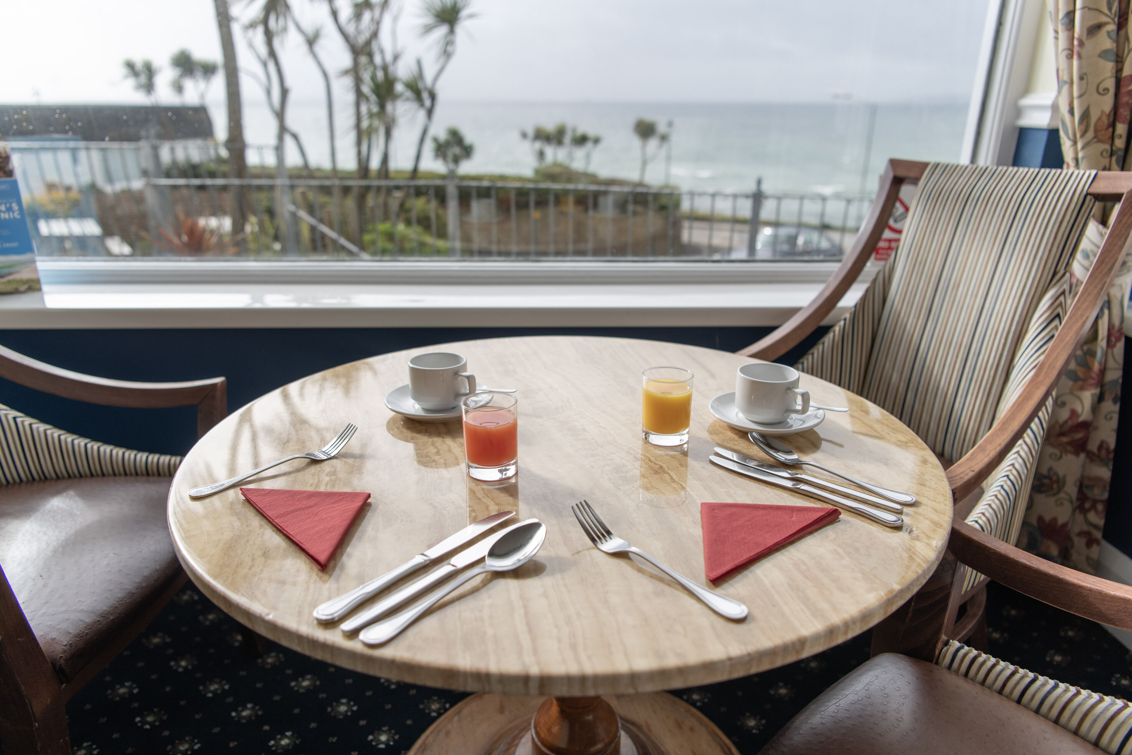 Une table ronde en bois, offrant une vue sur l’océan, agrémentée de couverts et de serviettes, et sur laquelle sont posés deux verres remplis de jus d’orange.