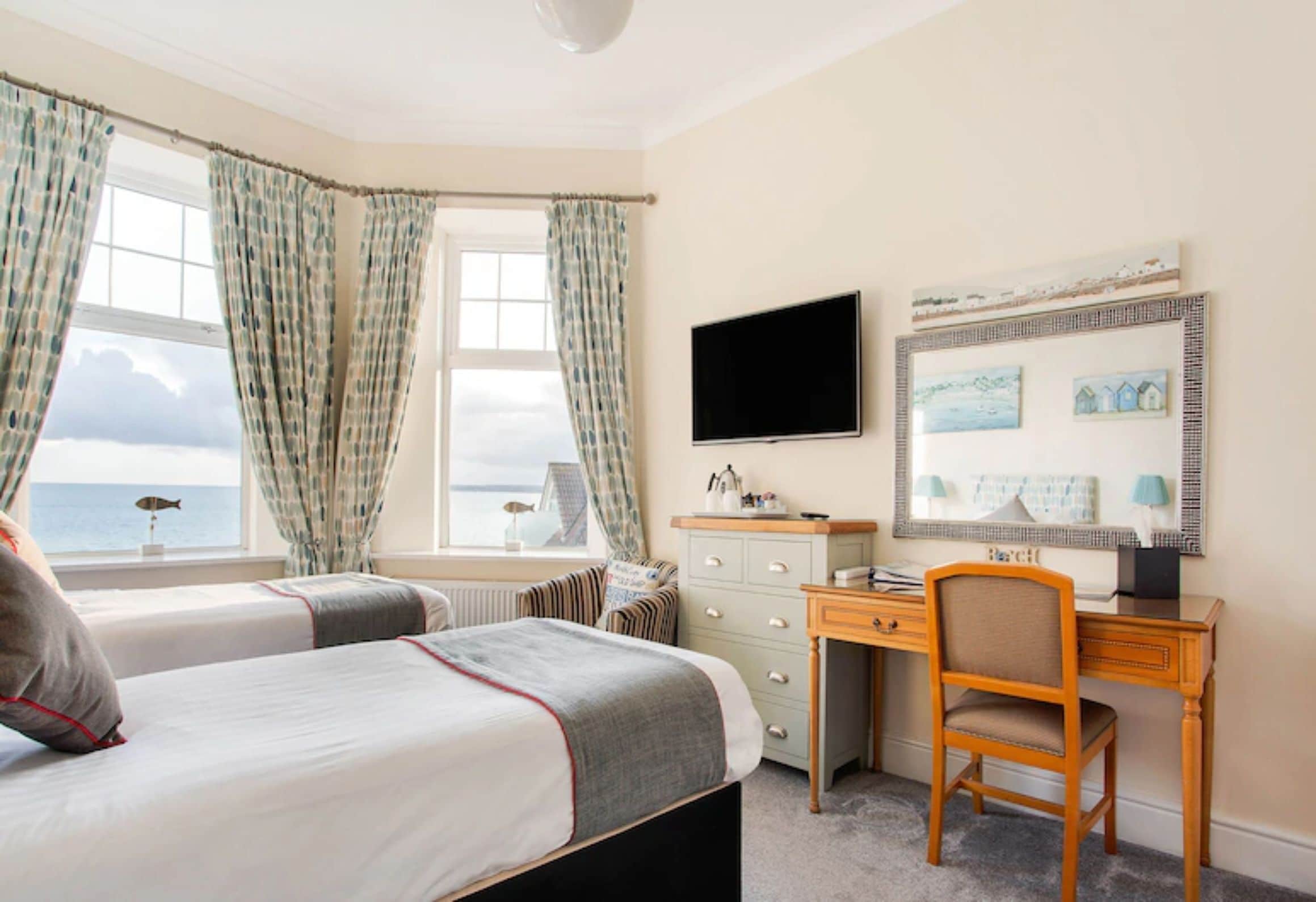 A hotel room with two beds, a desk, a chair, a television, and a view of the sea.