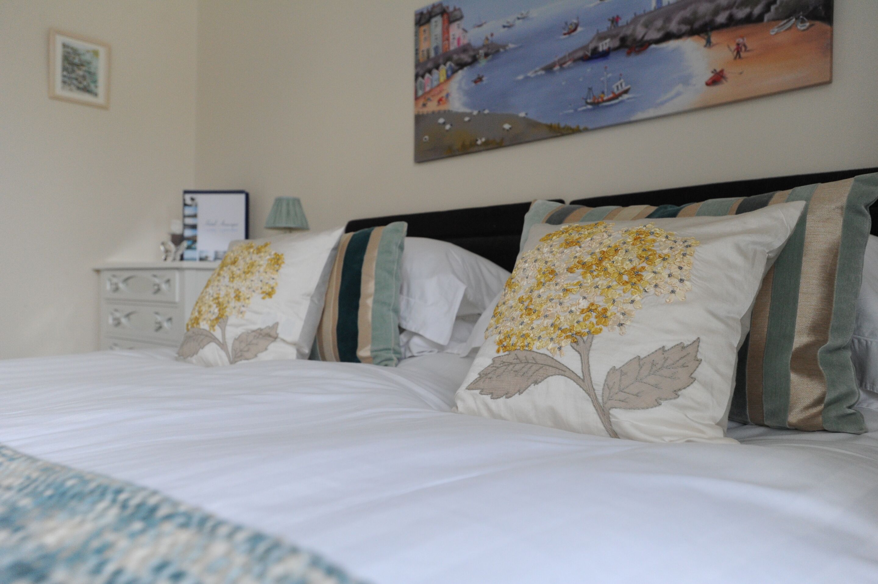 A neatly made bed with floral pillows, a bedside table with a lamp, and a painting of a coastal scene on the wall.