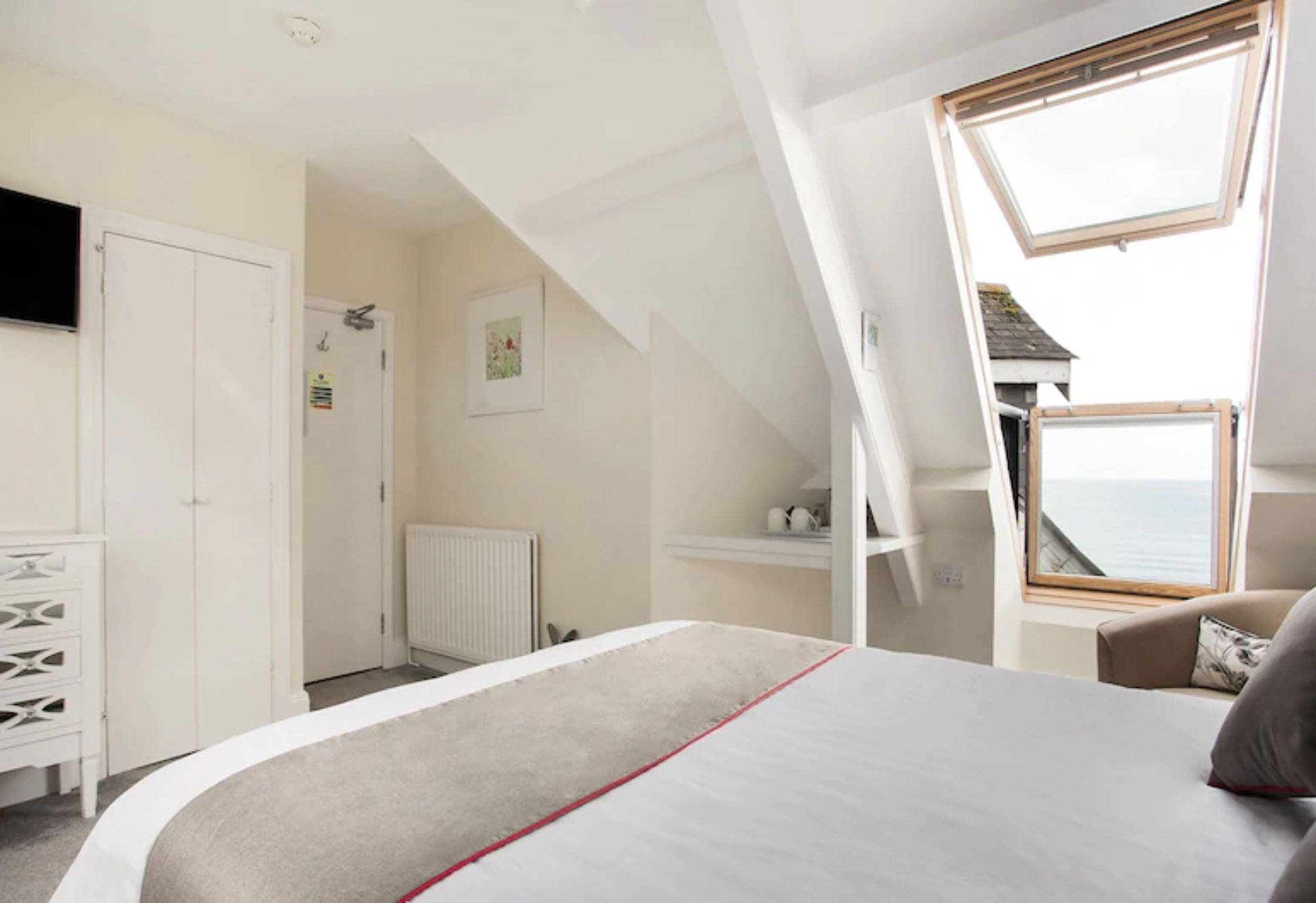 A bedroom with a bed, a dresser, a radiator, a window, and a view of the sea.