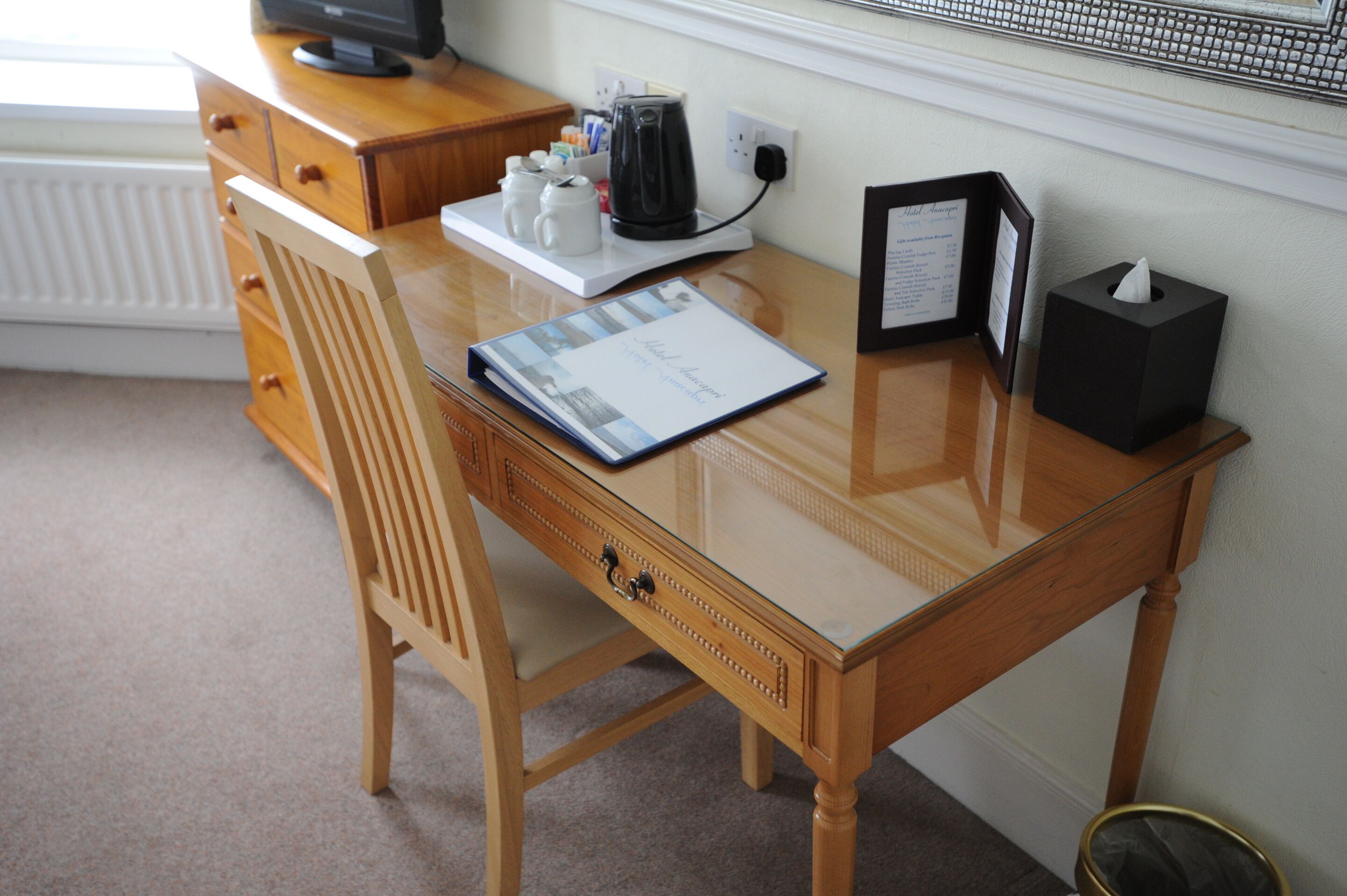 A wooden desk with a glass top, a coffee maker, a framed menu, and a tissue box.