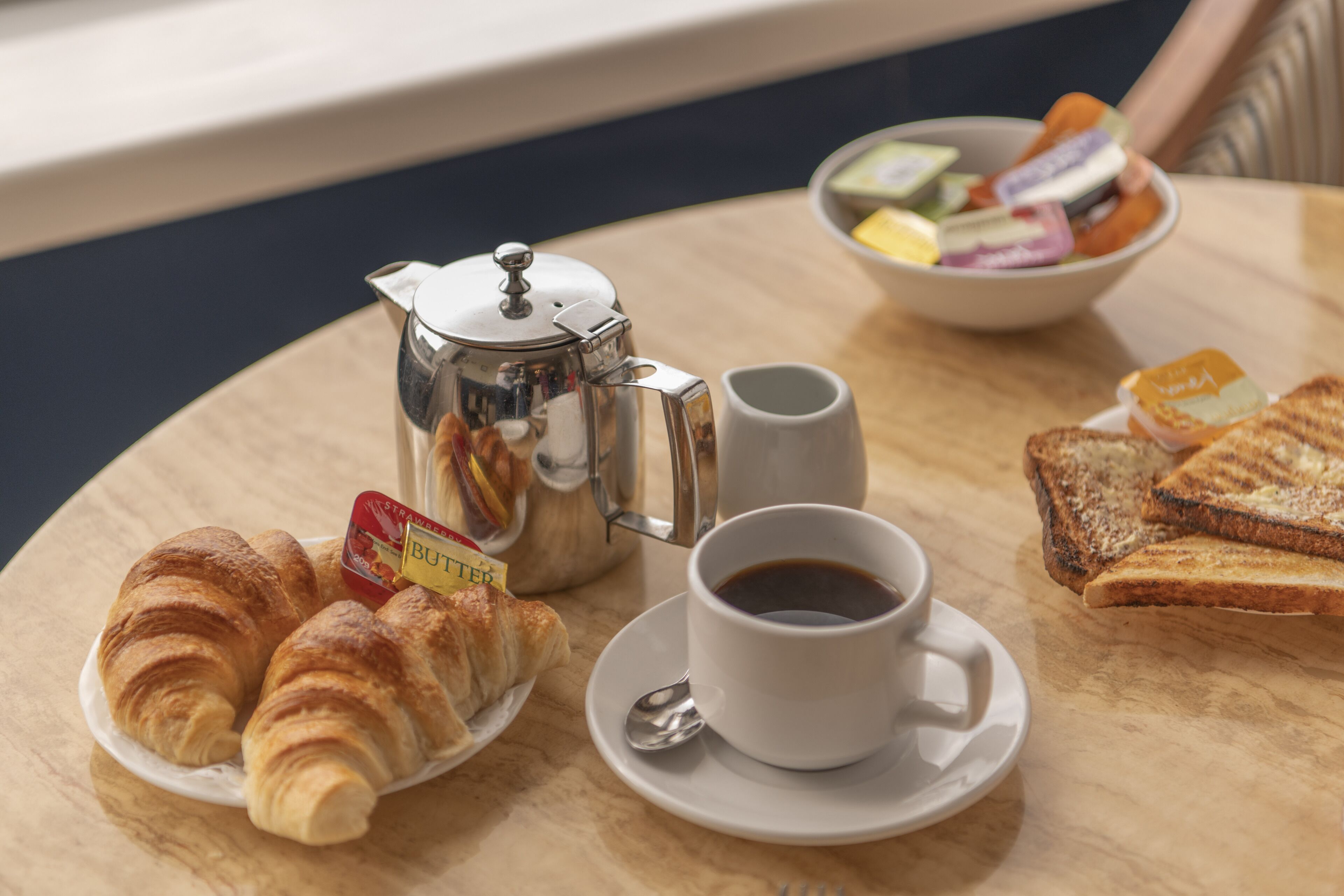 A breakfast setting with croissants, toast, a cup of coffee, a teapot, and a bowl of assorted fruits and yogurts.