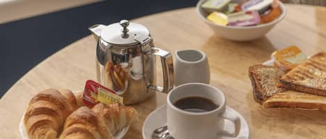 A breakfast setting with croissants, toast, a cup of coffee, a teapot, and a bowl of assorted fruits and yogurts.