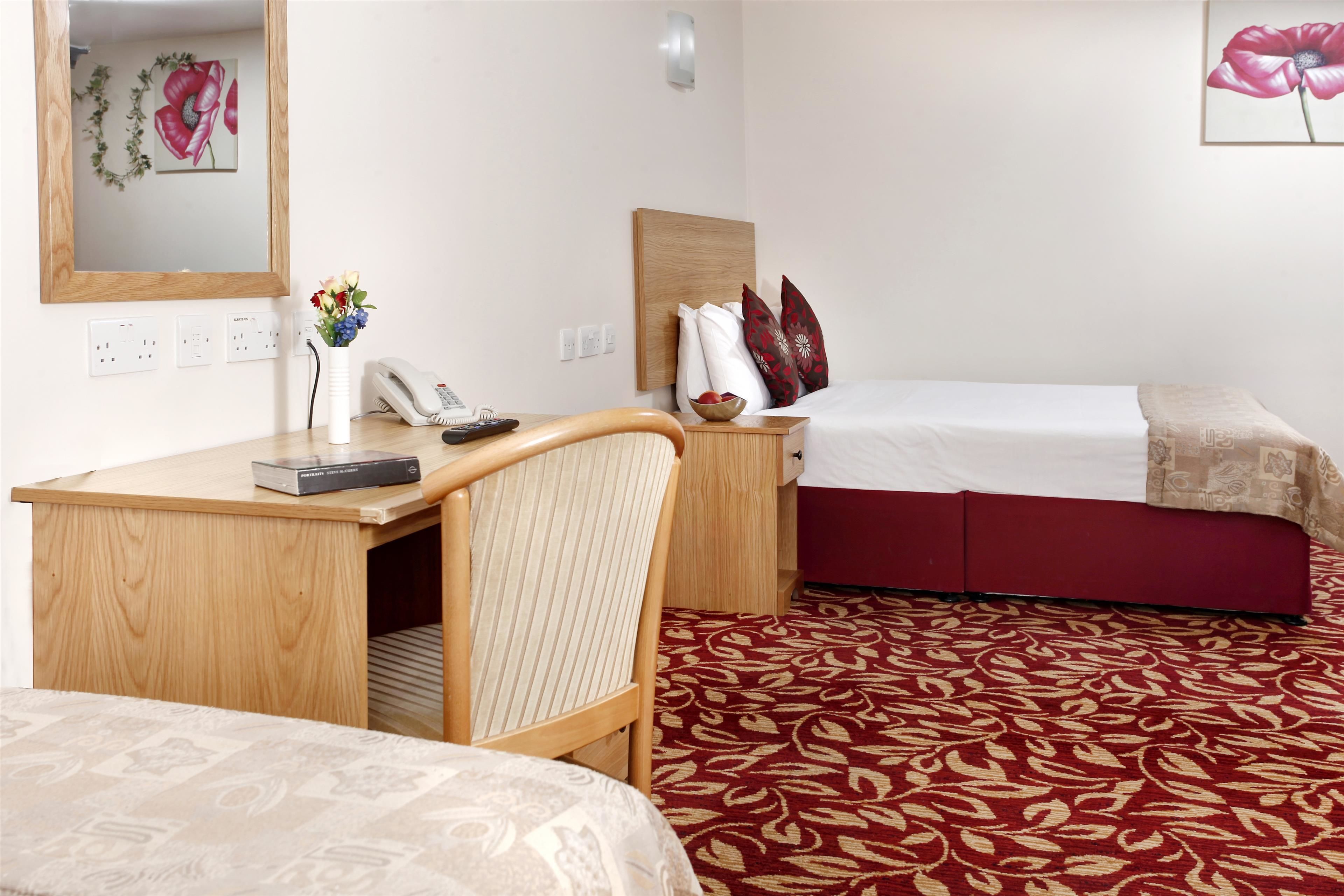 Ett hotellrum med ett skrivbord i trä, en stol, en säng med ett rött och vitt sängöverkast och en mönstrad matta.