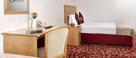 Habitación de hotel con un escritorio de madera, una silla, una cama con colcha roja y blanca, y una alfombra estampada.