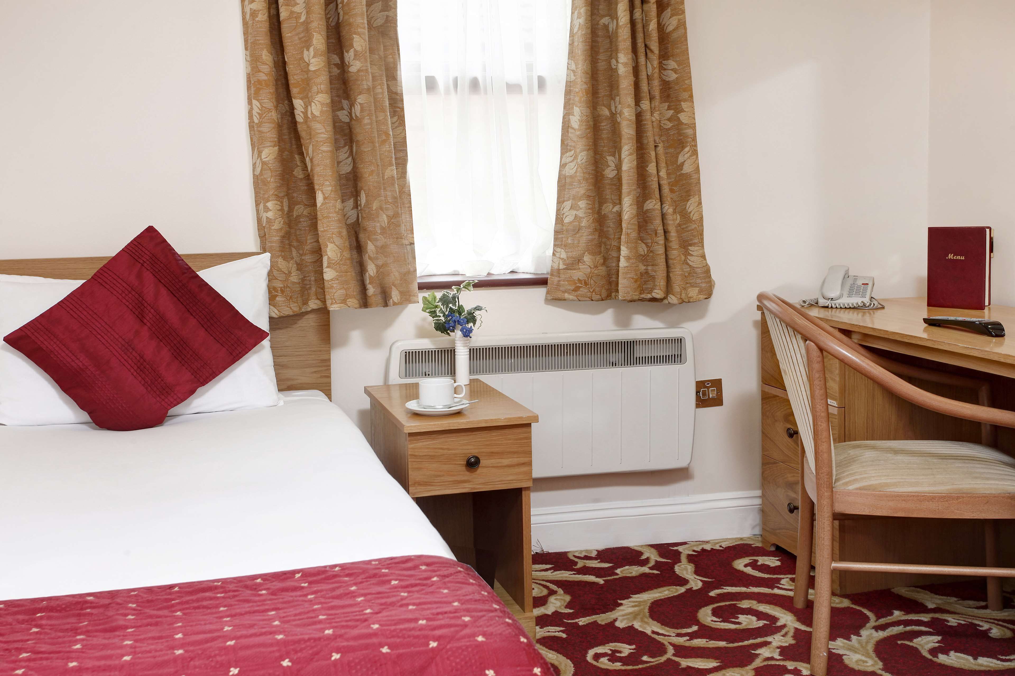 A hotel room with a bed, a desk, a chair, a radiator, and a window with curtains.