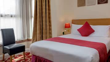 A hotel room with a bed, a chair, a nightstand with a lamp, and a window with curtains.