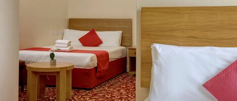 A hotel room with a wooden headboard, a red and white bedspread, a small round table with a vase, and a red flower painting on the wall.