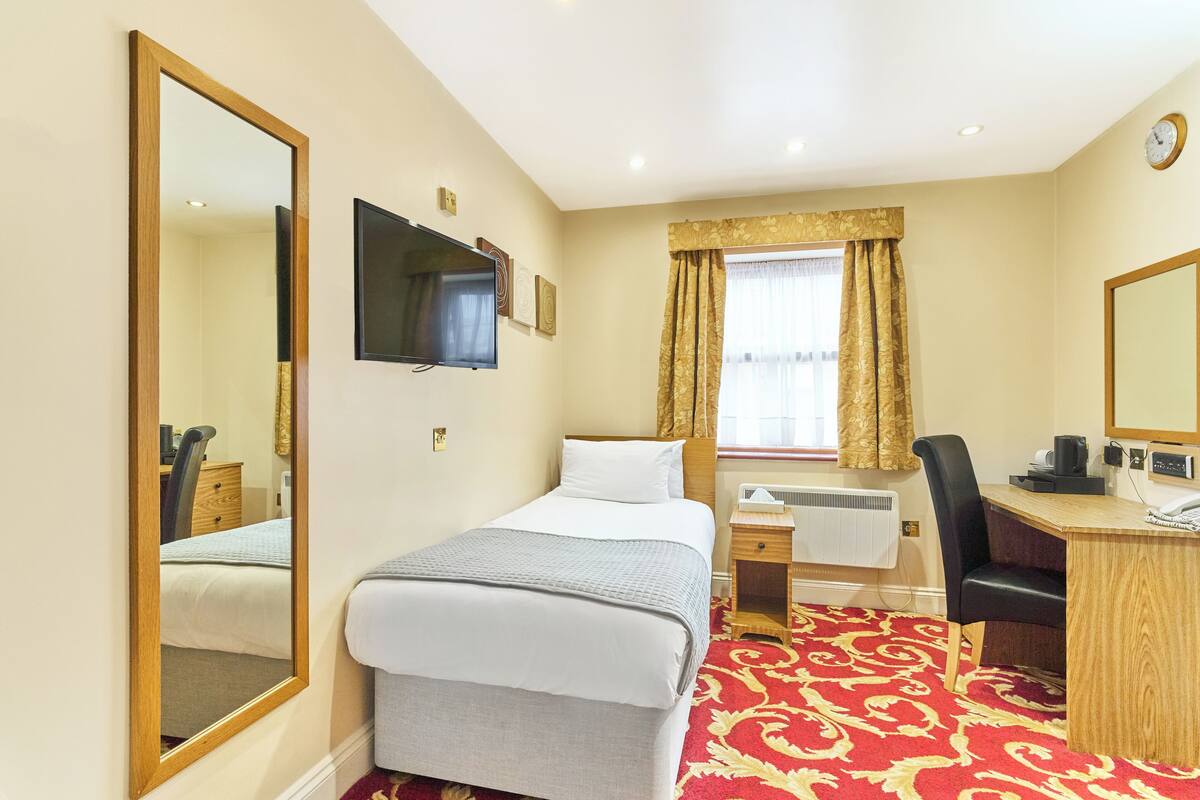 A hotel room with a bed, a desk, a chair, a TV, a window with curtains, and a clock on the wall.