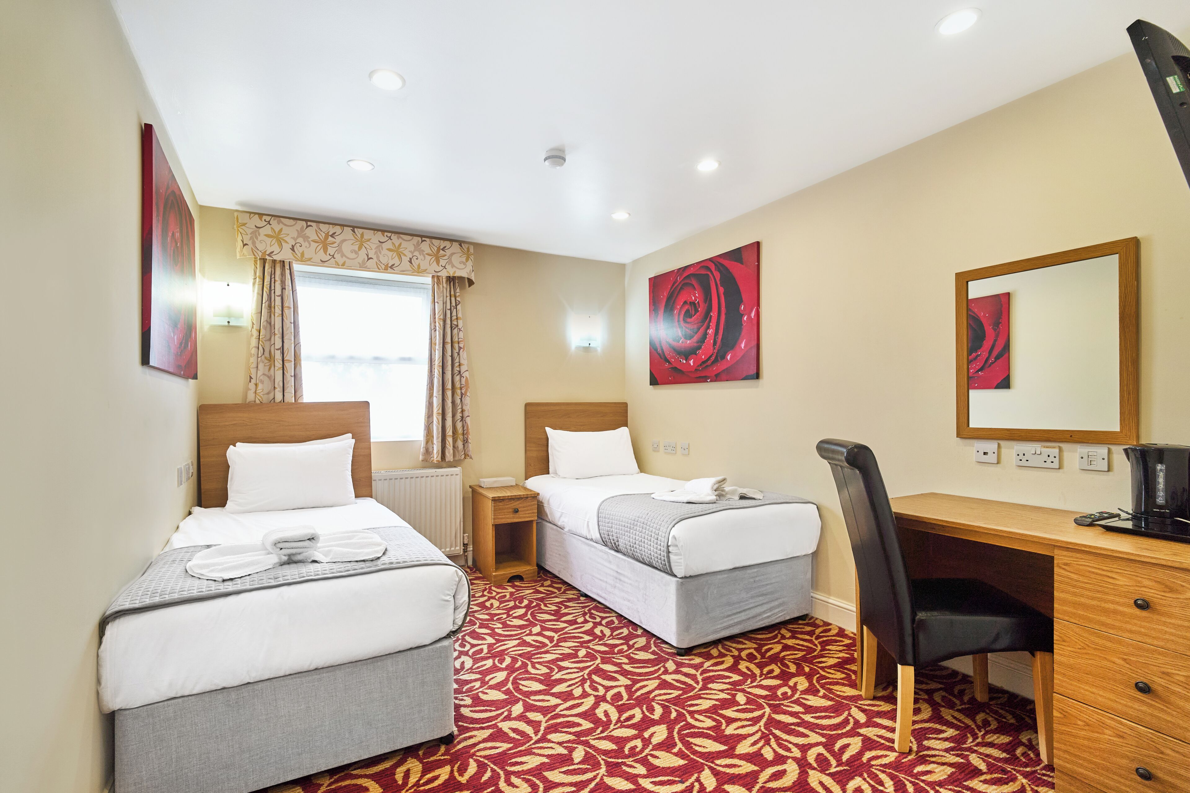 Habitación de hotel con dos camas, un escritorio, una silla, un ventanal con cortinas y obras de arte en las paredes.