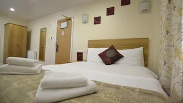 Una cama bien tendida con sábanas blancas y un cojín decorativo, un armario de madera y una lámpara fijada a la pared.
