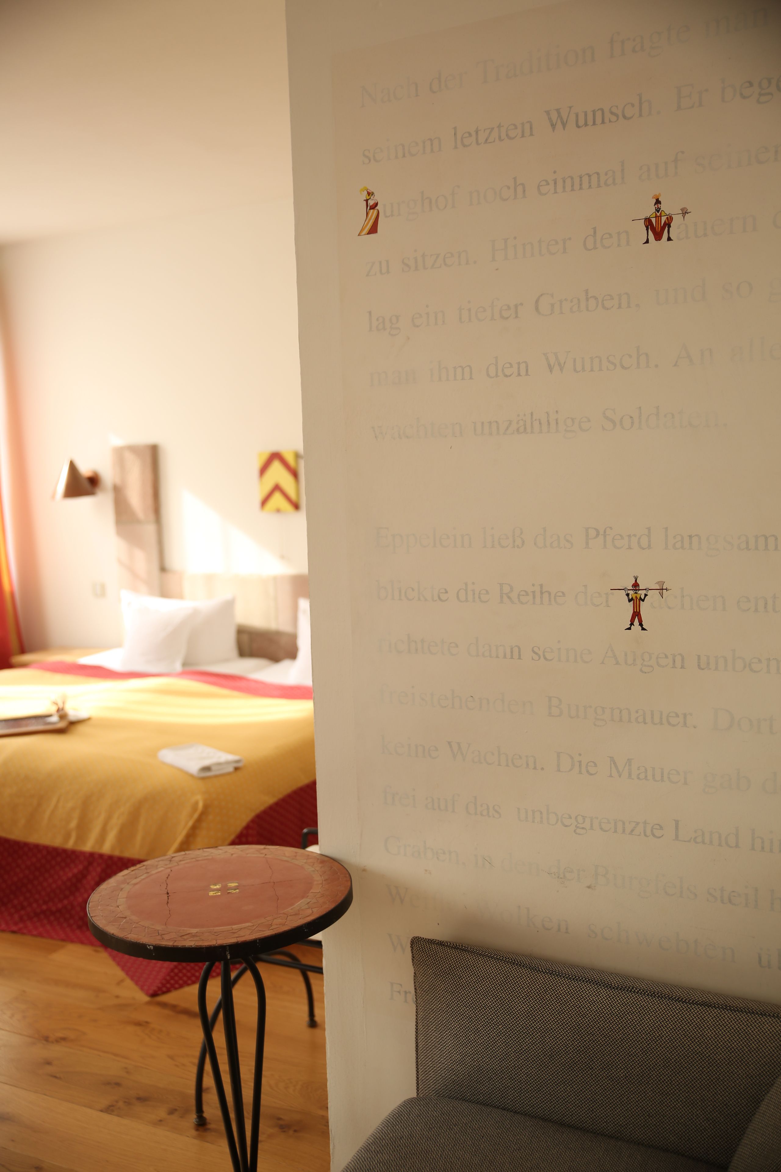 Quarto de hotel com uma cama, uma mesa redonda pequena e uma parede com texto e ilustrações.
