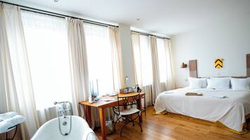 Habitación de hotel con suelo de madera, bañera, escritorio con silla y cama con ropa de cama blanca.