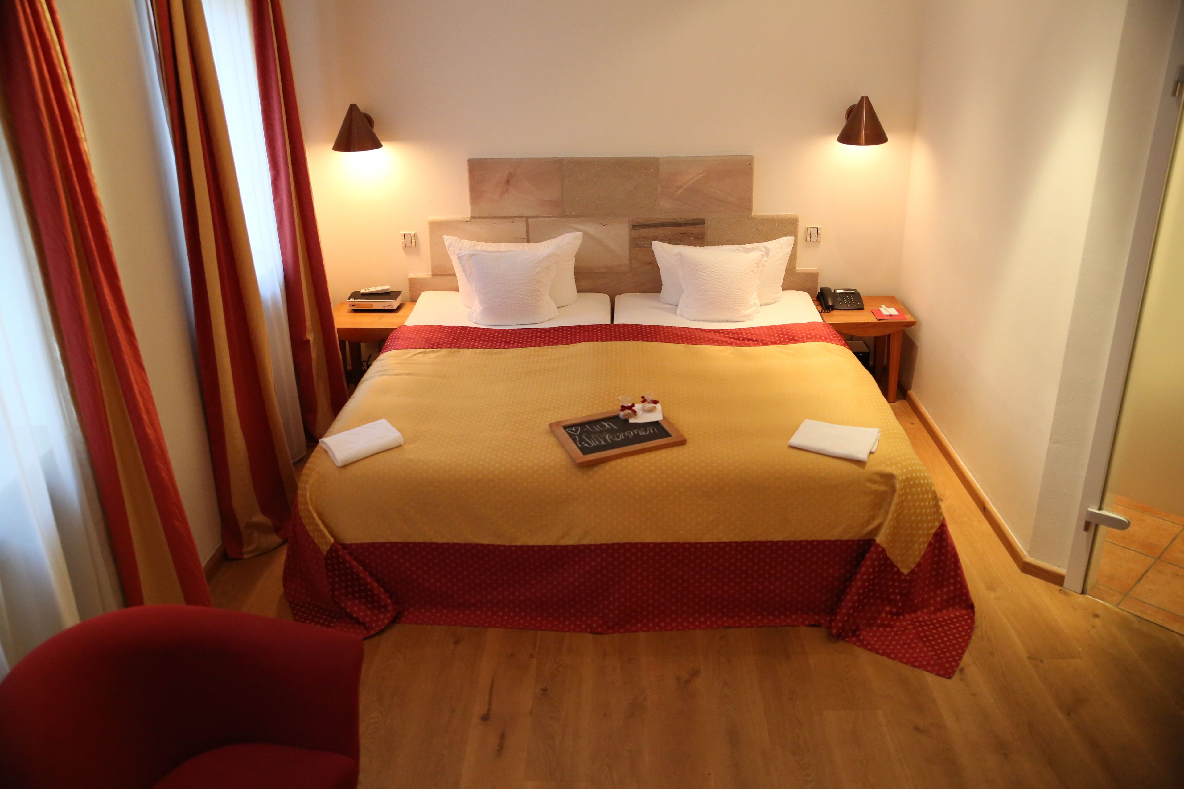 A hotel room with a large bed, two bedside tables, a nightstand, a red chair, and a wooden floor.