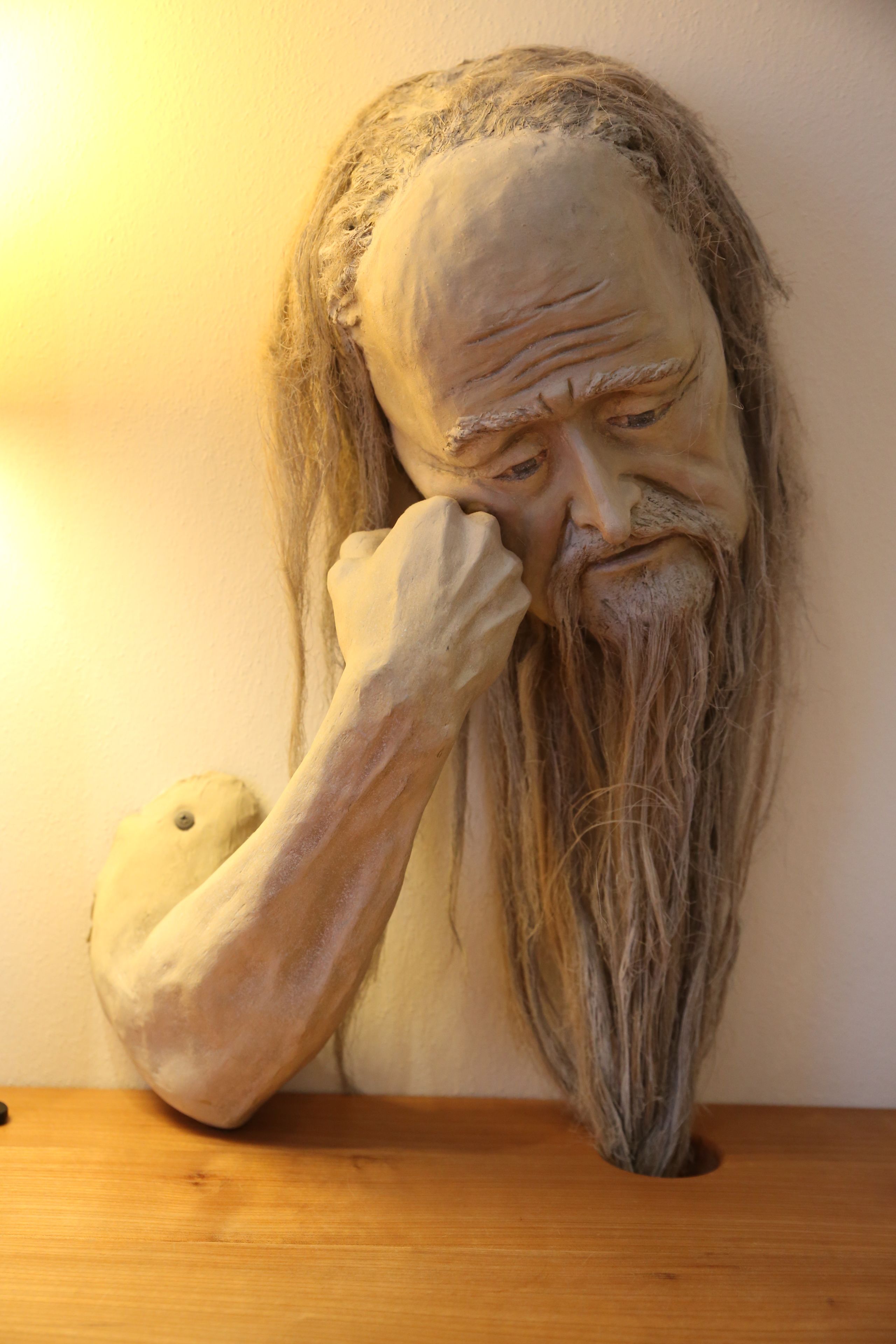 Una scultura raffigura una persona con capelli lunghi e barba, posata su una superficie in legno di fronte a un muro spoglio.