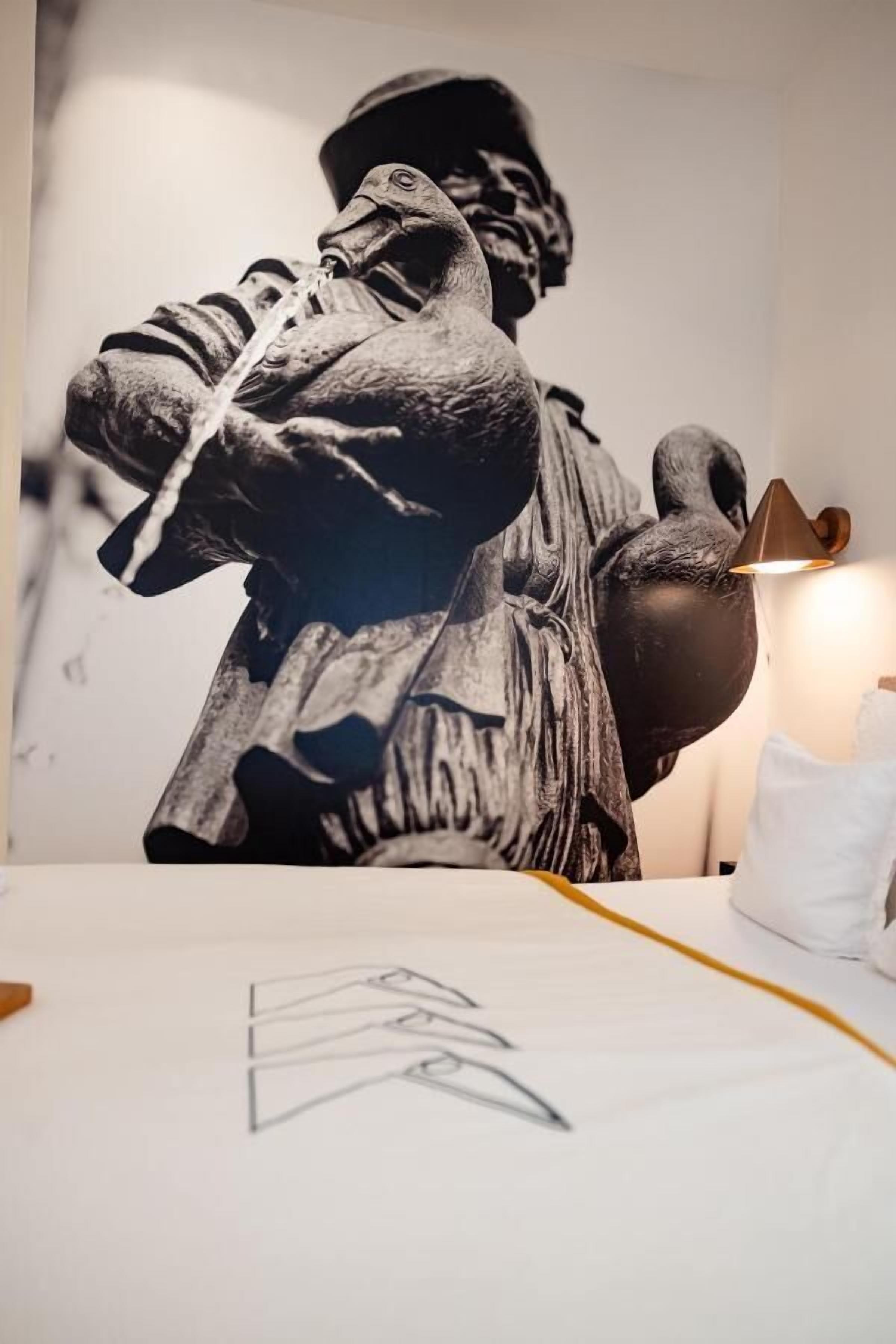 Una habitación con una estatua grande en blanco y negro de una persona sosteniendo una cuerda, una lámpara y un almohadón blanco sobre una cama.