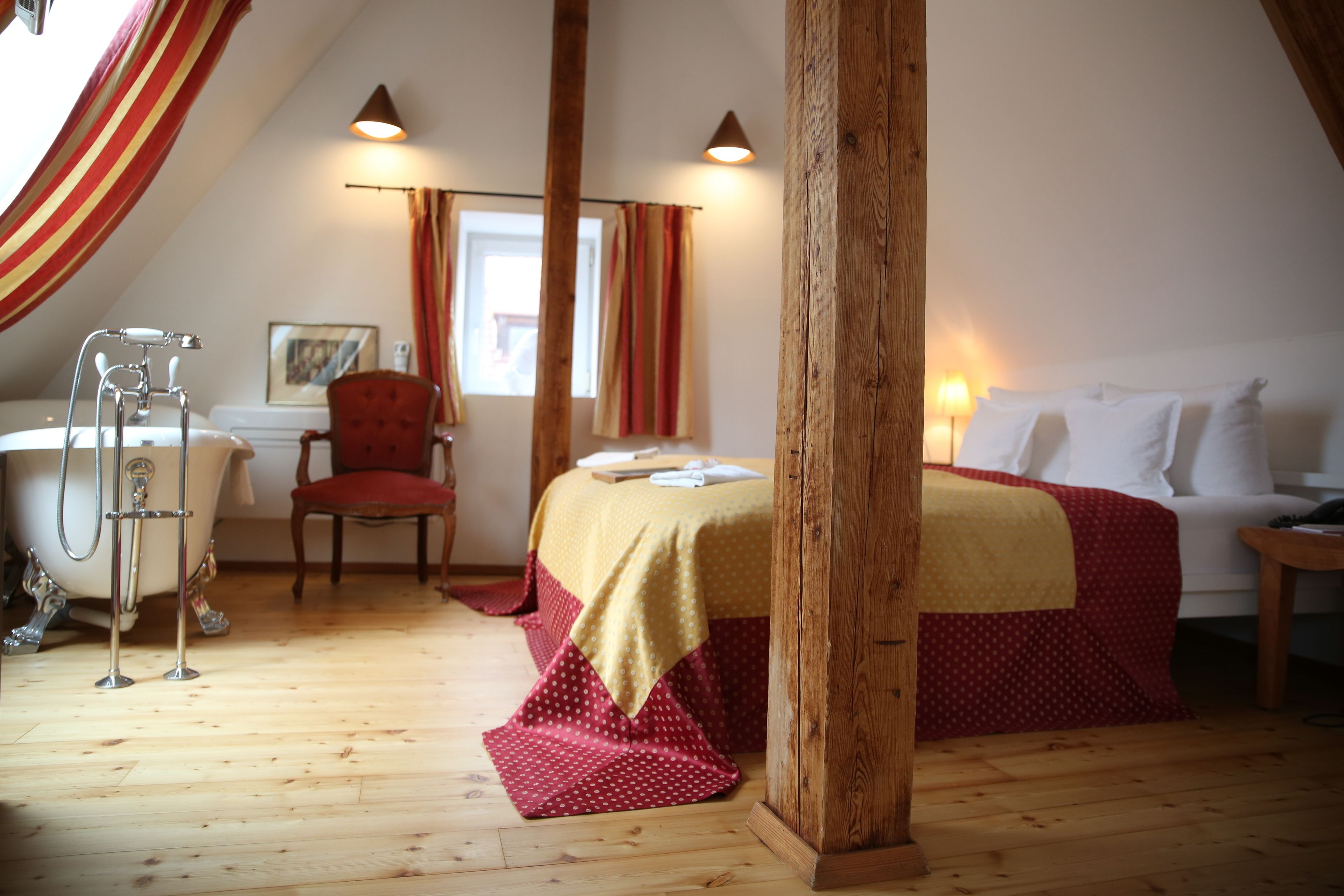Quarto com piso de madeira, cama com colcha vermelha e amarela, cadeira vermelha, sofá branco, mesa de cabeceira de madeira e viga de madeira no teto.