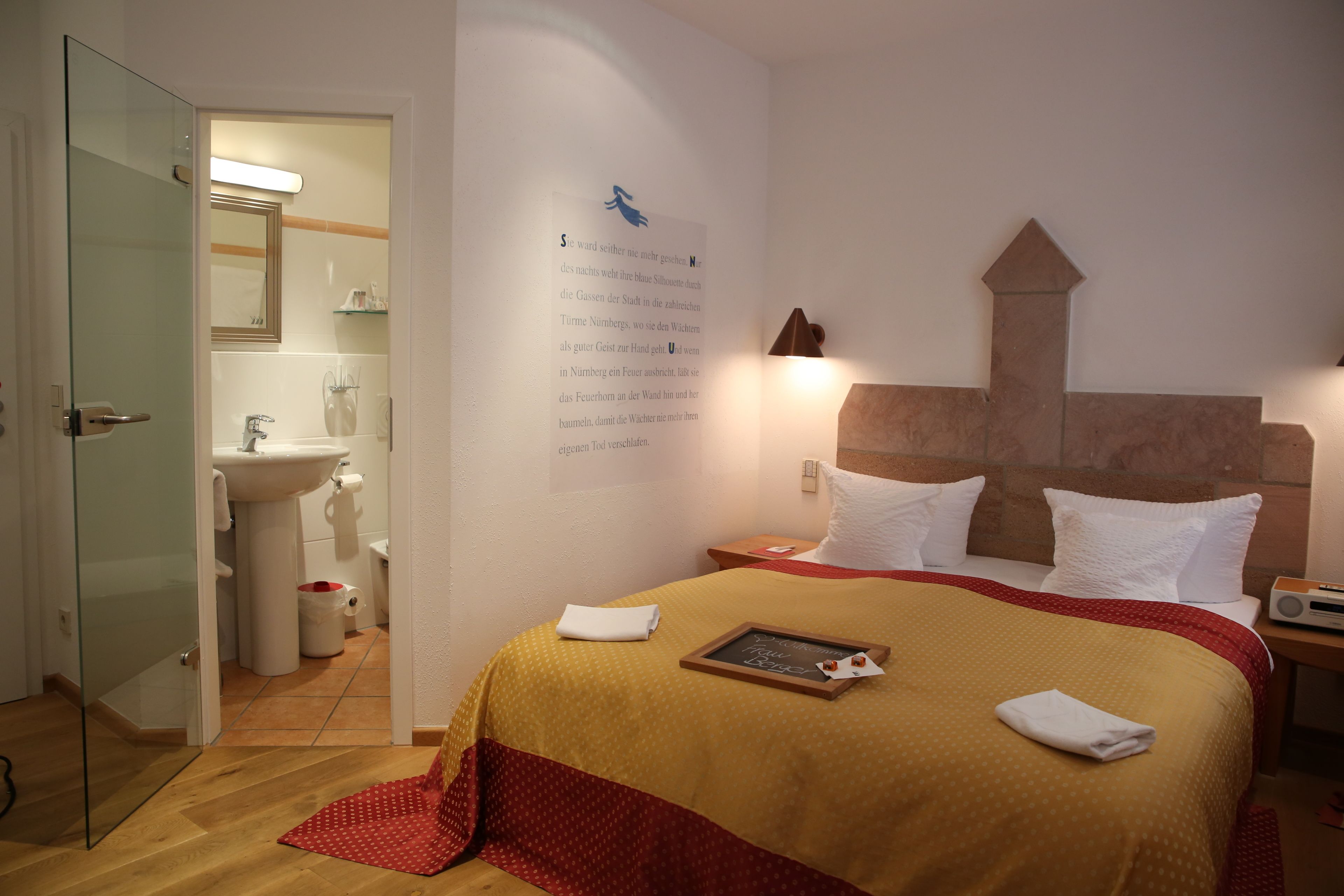 A hotel room with a bed, a bedside table, a nightstand, a wall-mounted lamp, a framed picture, and a bathroom visible through an open door.