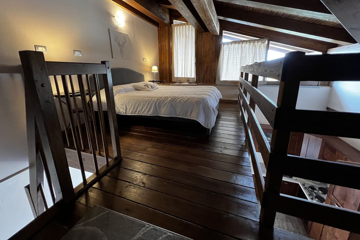 A wooden-floored bedroom with a bed, a nightstand, a wall-mounted lamp, and a window with curtains.