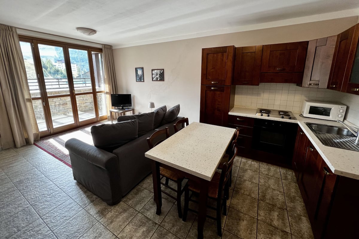 A compact living space with a sofa, a dining table, a kitchen area with a microwave, and a large window.