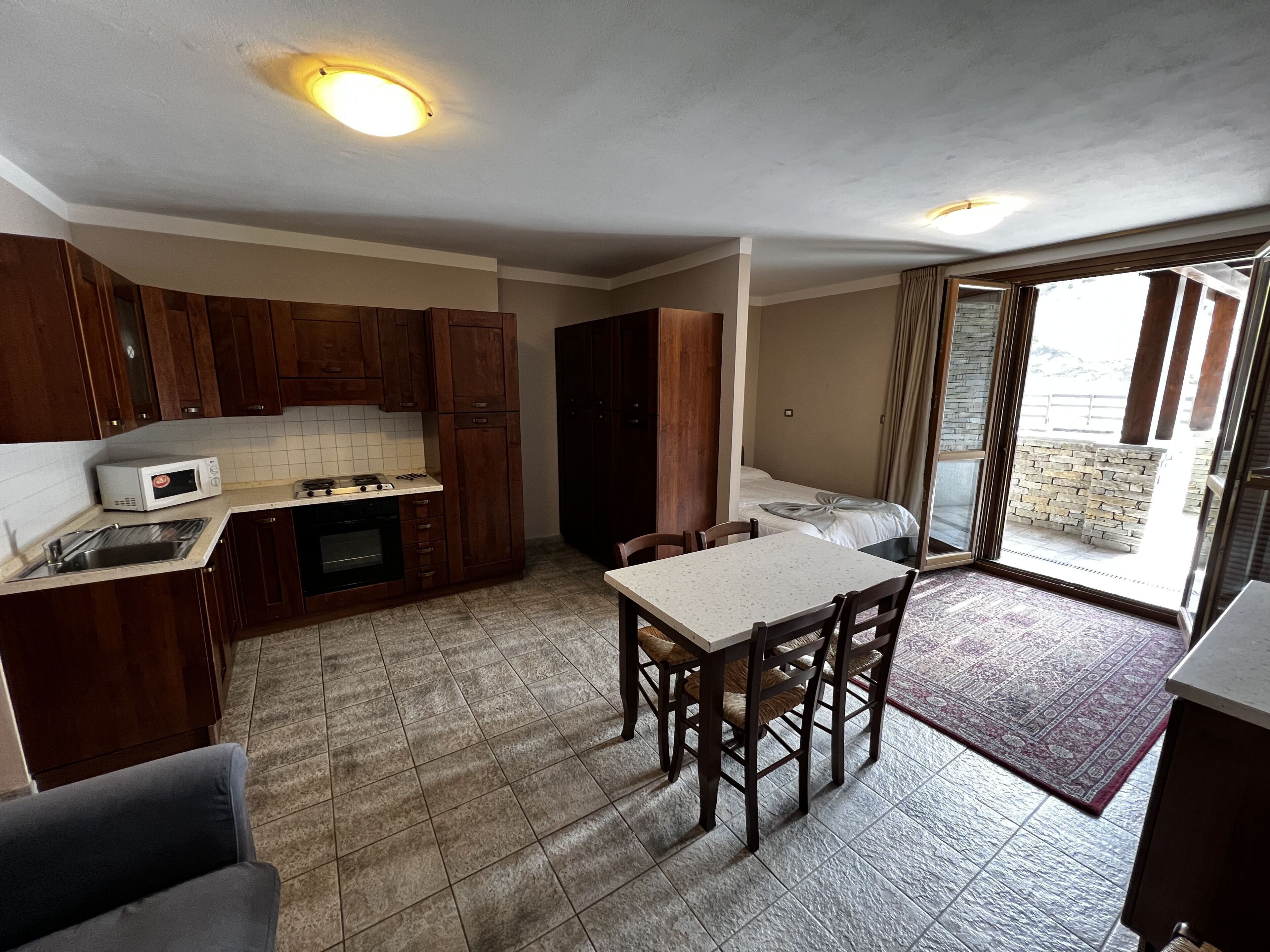 A compact kitchen with a microwave, stove, and sink, a dining table with chairs, and a view of a balcony with stone walls.