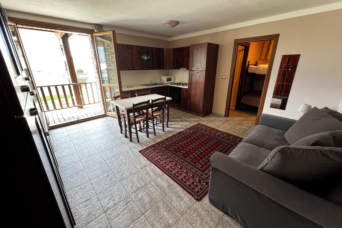 A spacious living area with a sofa, a dining table with chairs, a kitchen with wooden cabinets, and a sliding glass door leading to an outdoor balcony.