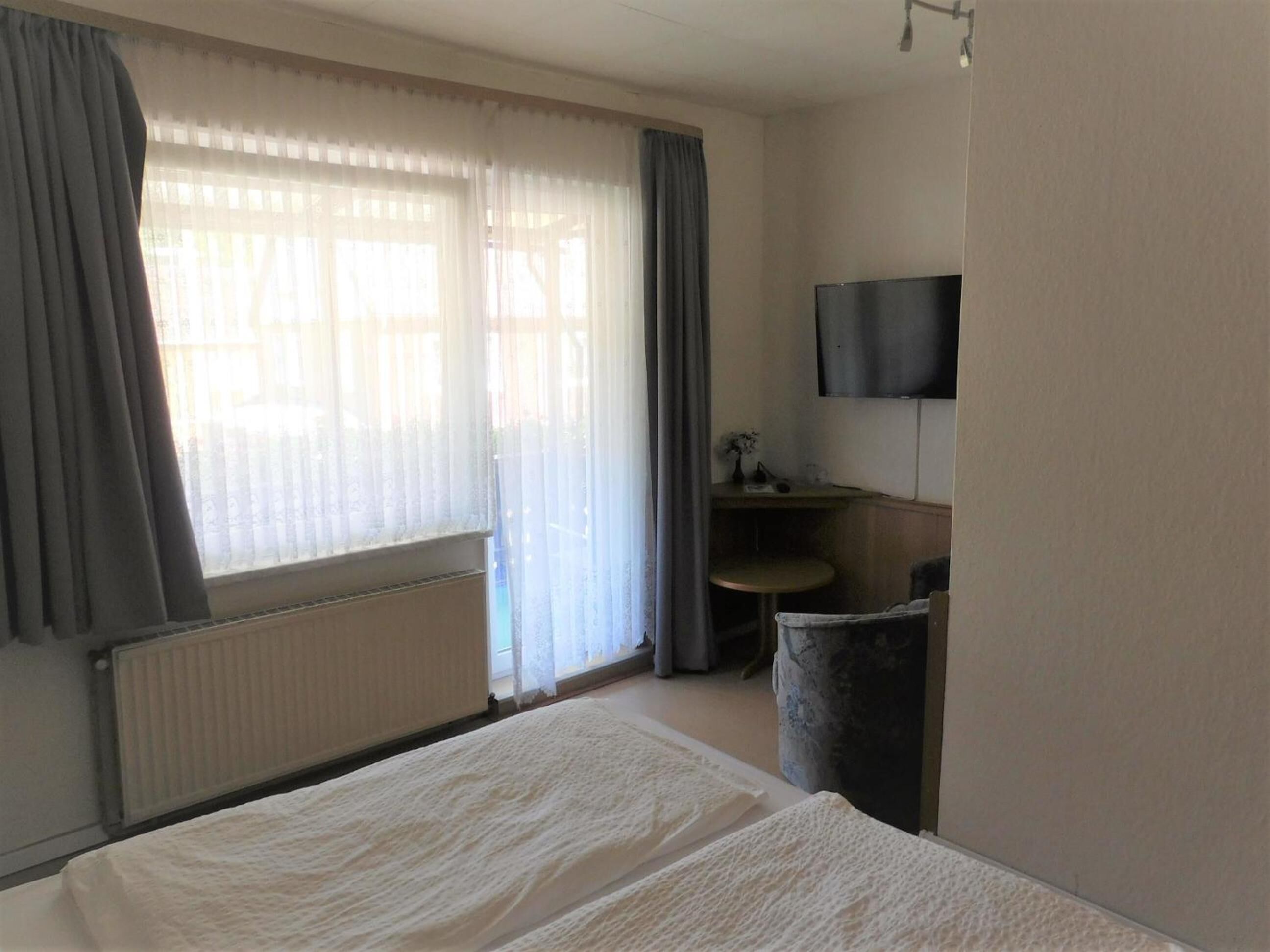 A hotel room with a bed, a desk, a chair, a radiator, a TV mounted on the wall, and a window with curtains.